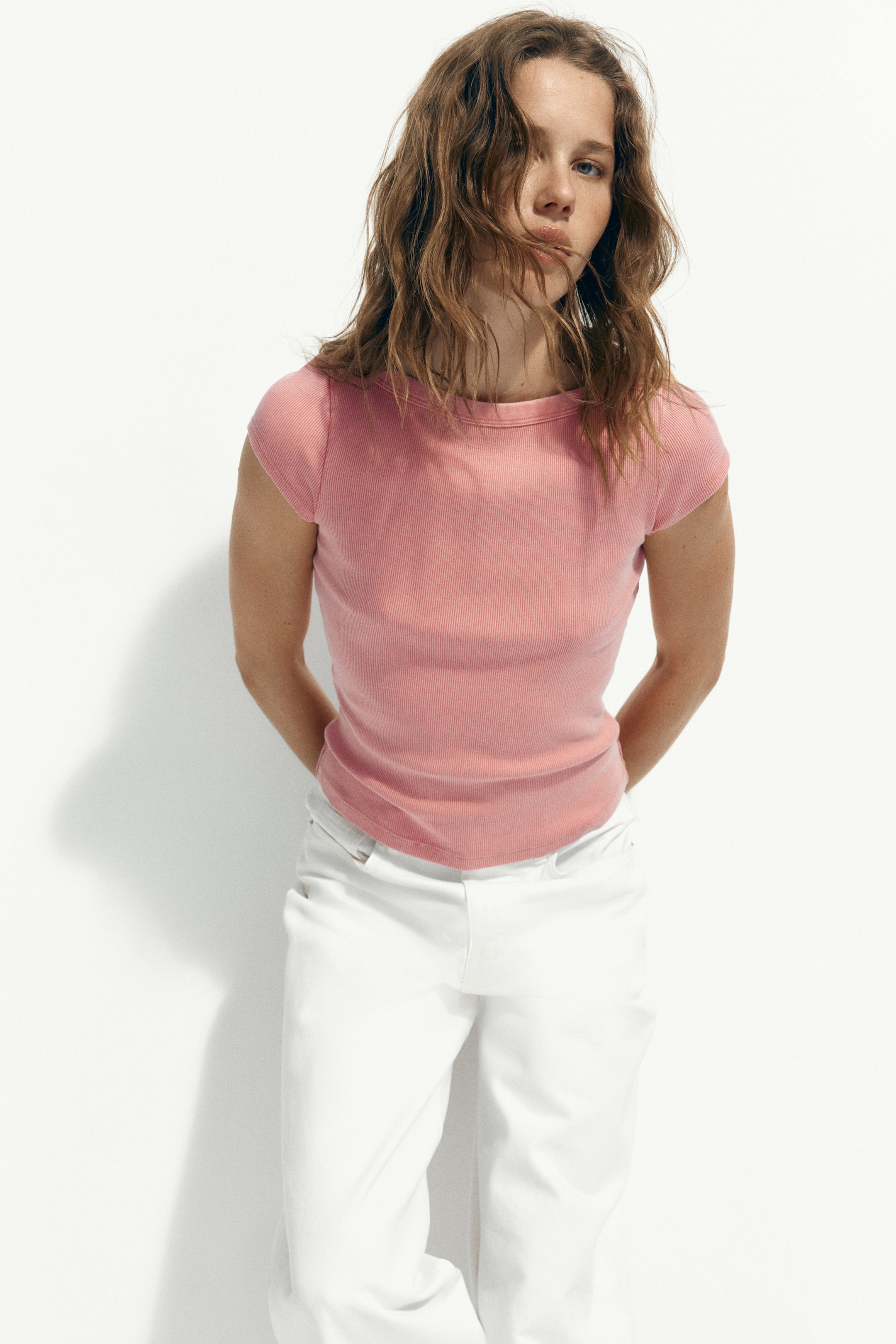 拡大画像を表示: ピンクのリブTシャツを着用しており、クルーネック、ショートキャップスリーブ、肩に見える縫い目のディテールが特徴です。スリムフィットとテクスチャードニットを披露しています。