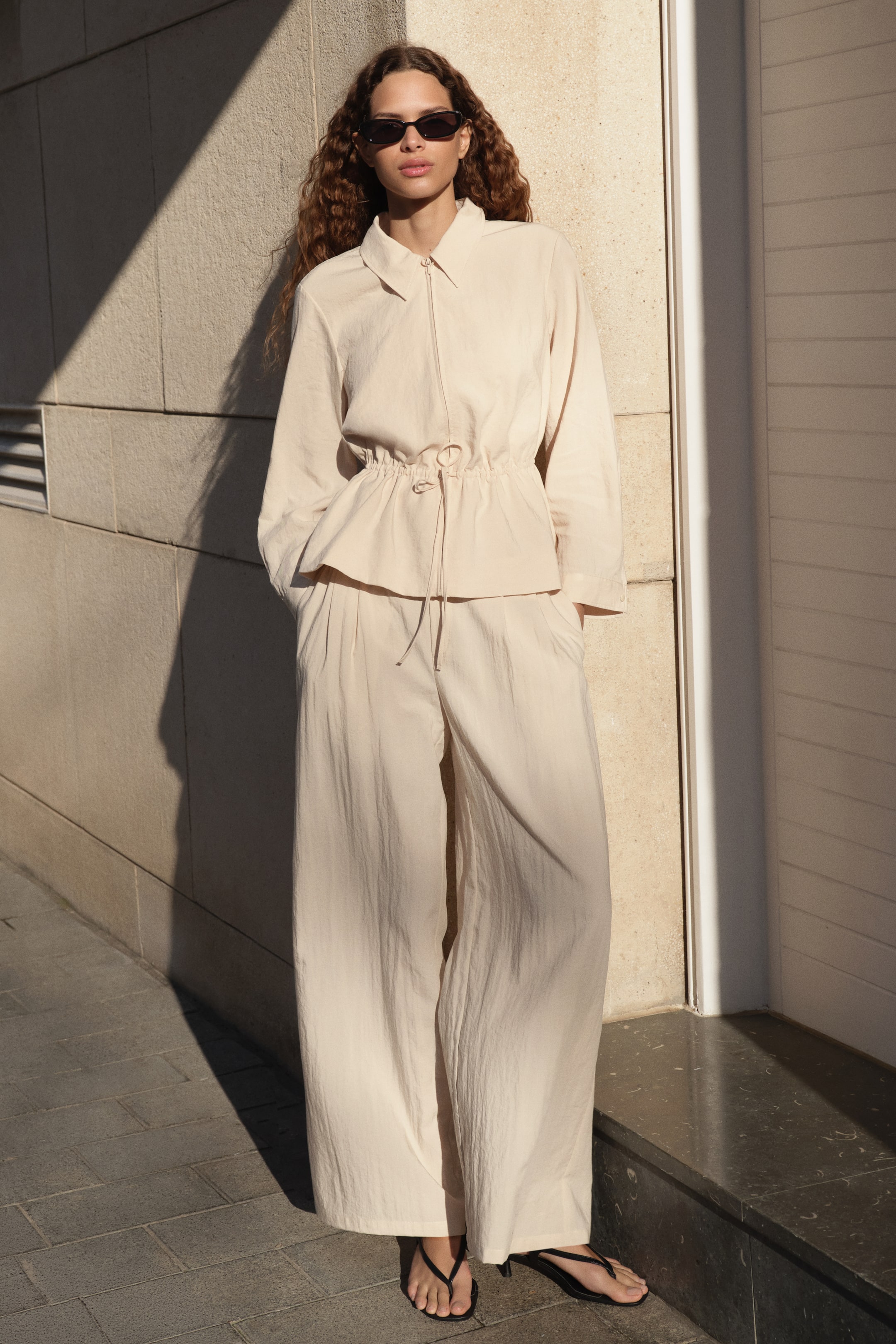View larger image: A light beige two-piece set crafted from crinkled fabric, showcasing a collared, long-sleeved jacket with a front zipper and a drawstring waist, forming a peplum hem. It is complemented by matching wide-leg trousers.
