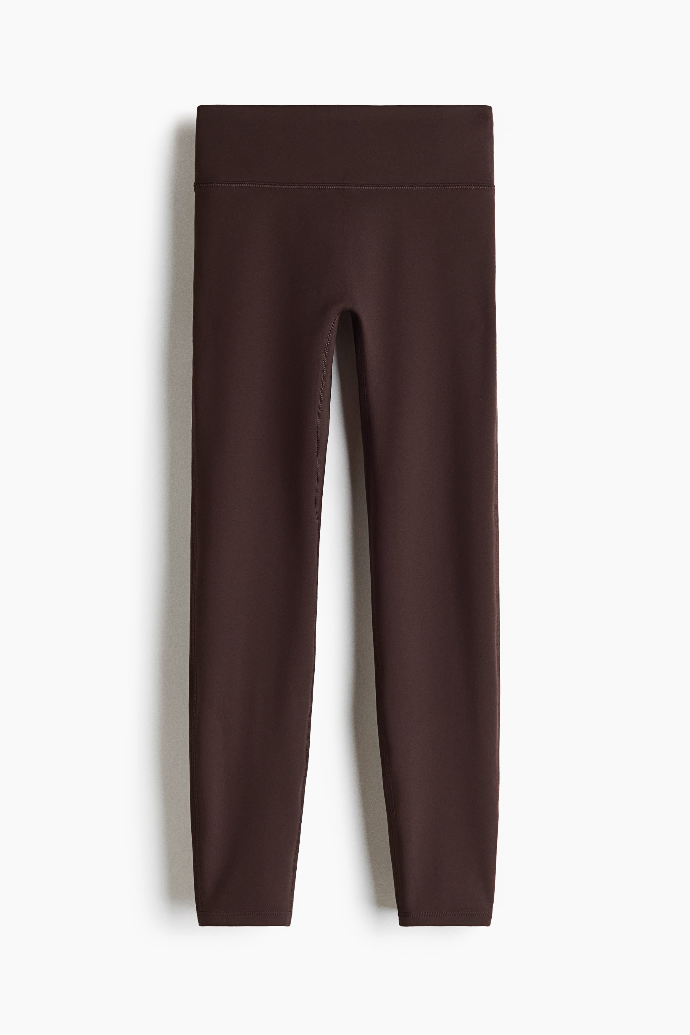 Een sportieve outfit uit drie delen wordt van voren getoond, bestaande uit een crÃ¨mekleurig jack met rits en opstaande kraag, gedragen over een donkerbruine tanktop en bijpassende, hoog getailleerde leggings in volledige lengte. Het jack hangt open en accentueert het aansluitende silhouet van de donkerbruine stukken.