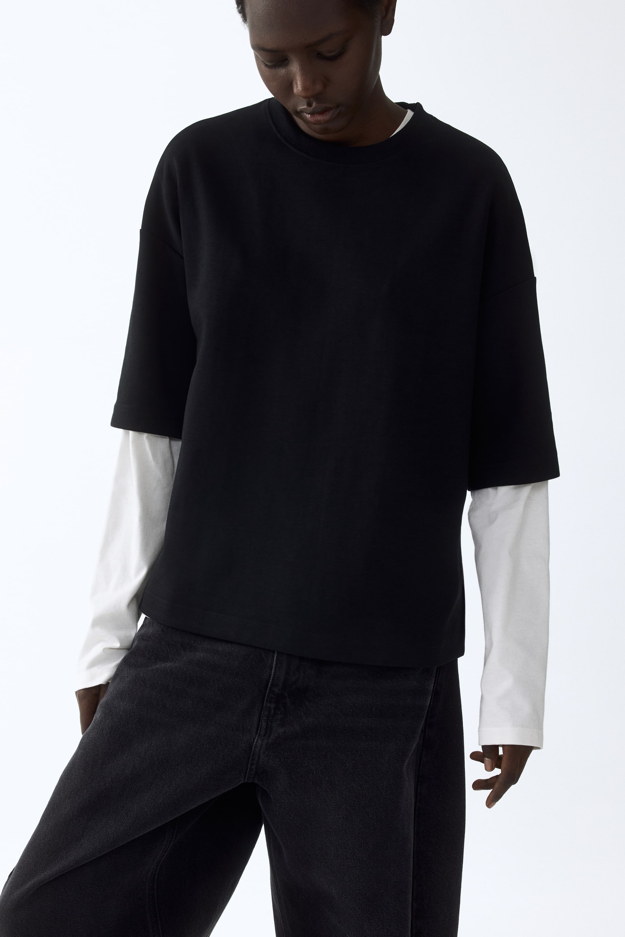 Ver imagem maior: A camisola preta de manga curta e gola redonda, com um corte descontraído, é apresentada num ângulo de frente e de perfil. Está sobreposta a uma camisa branca de manga comprida, visível na gola e nos punhos.