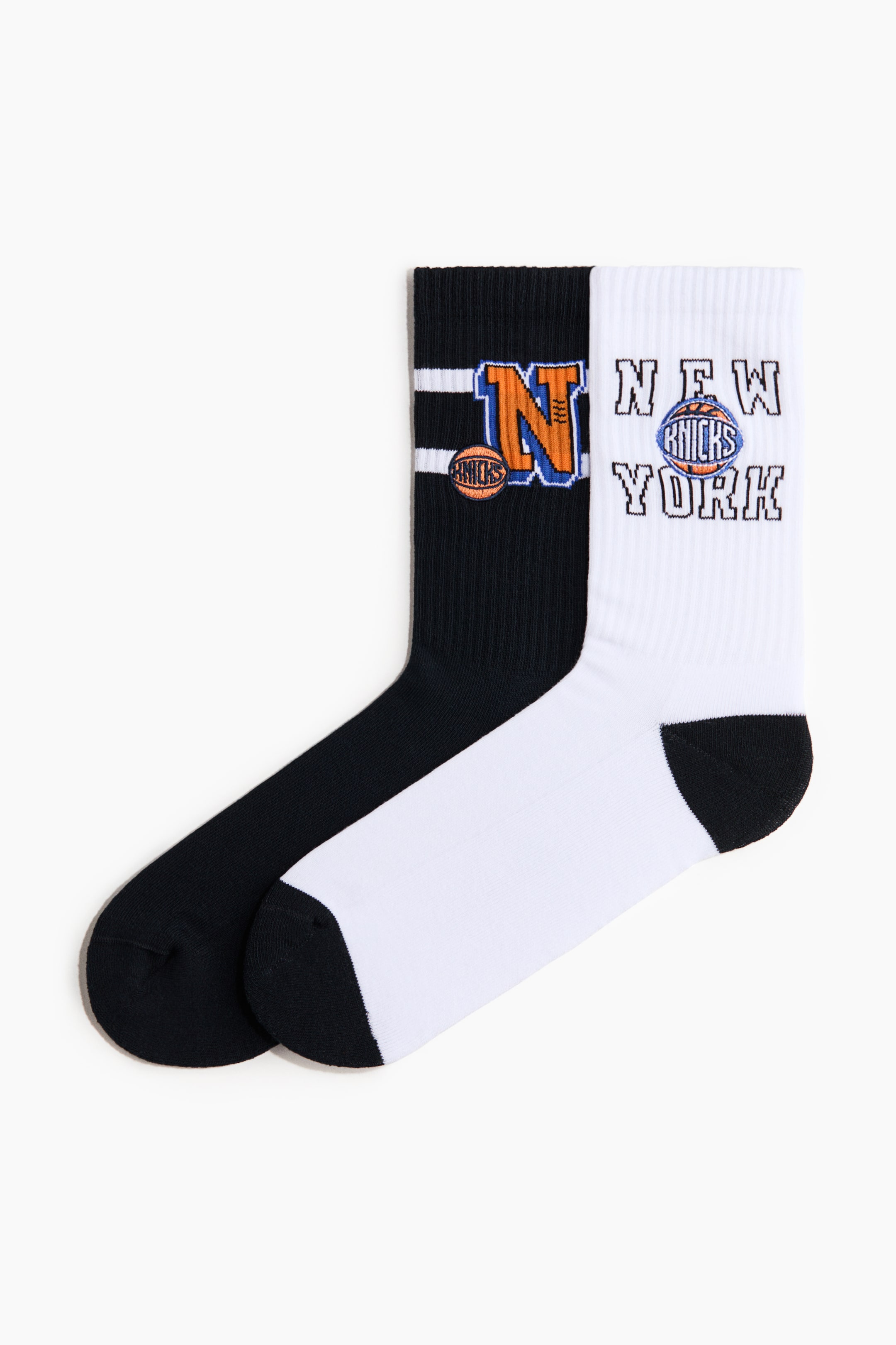 Visualizza l'immagine ingrandita: Un paio di calzini è disteso, mostrando un calzino nero e un calzino bianco con le parti frontali visibili. Il calzino nero presenta un grande motivo 'N' arancione e blu con un logo di basket e strisce bianche sul suo gambale a coste. Il calzino bianco mette in evidenza il testo 'NEW YORK' che fiancheggia un logo di basket con 'KNICKS' all'interno, sul suo gambale a coste. Entrambi i calzini presentano sezioni del tallone e della punta nere.