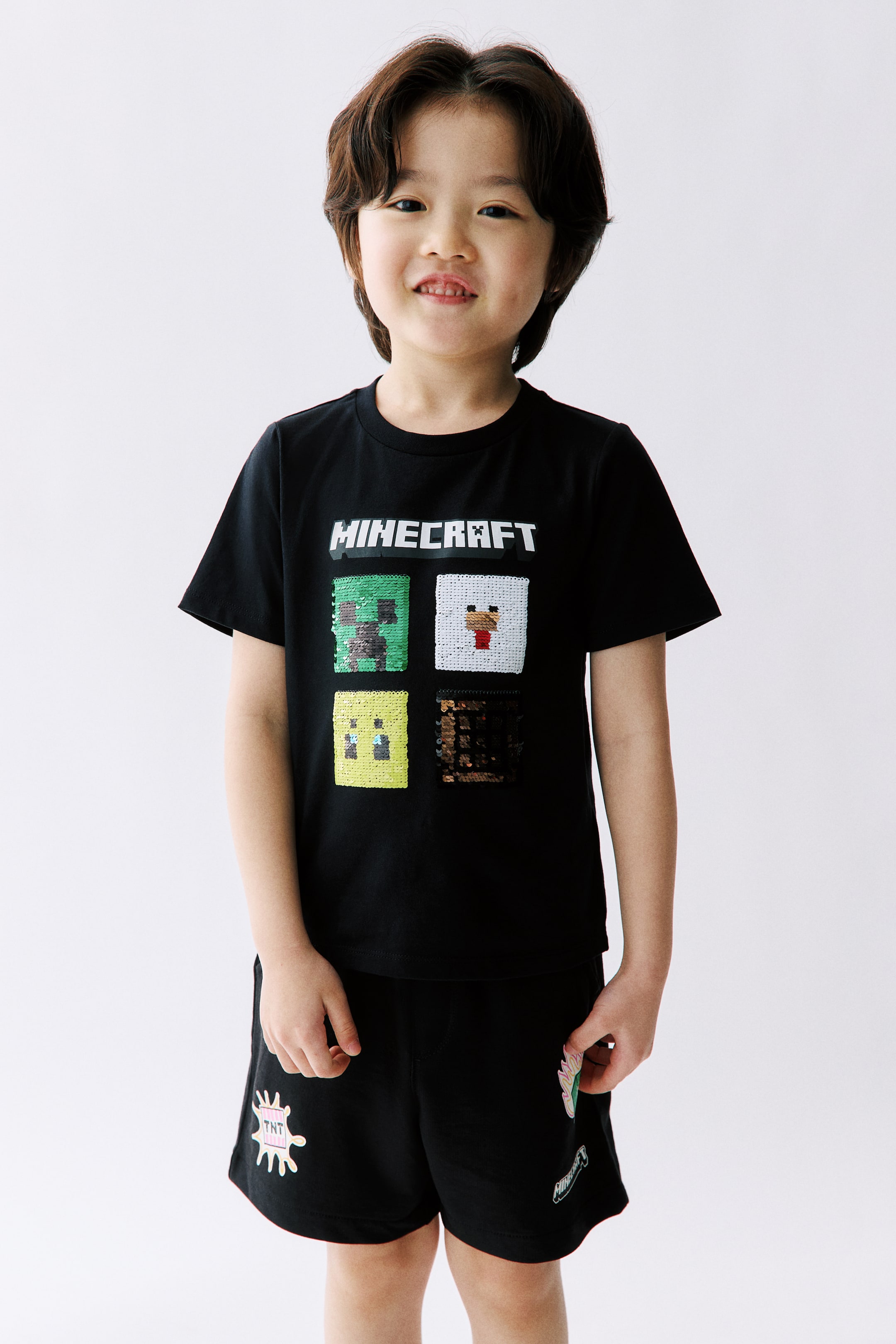 拡大画像を表示: モデルが着用しているのは、プリントコットンジャージー素材のTシャツで、丸いリブトリムのネックラインとストレートカットの裾が特徴です。ブラックのTシャツには、フロントに「MINECRAFT」ロゴと、4つのスパンコールで装飾されたピクセルキャラクターのスクエアが描かれています。