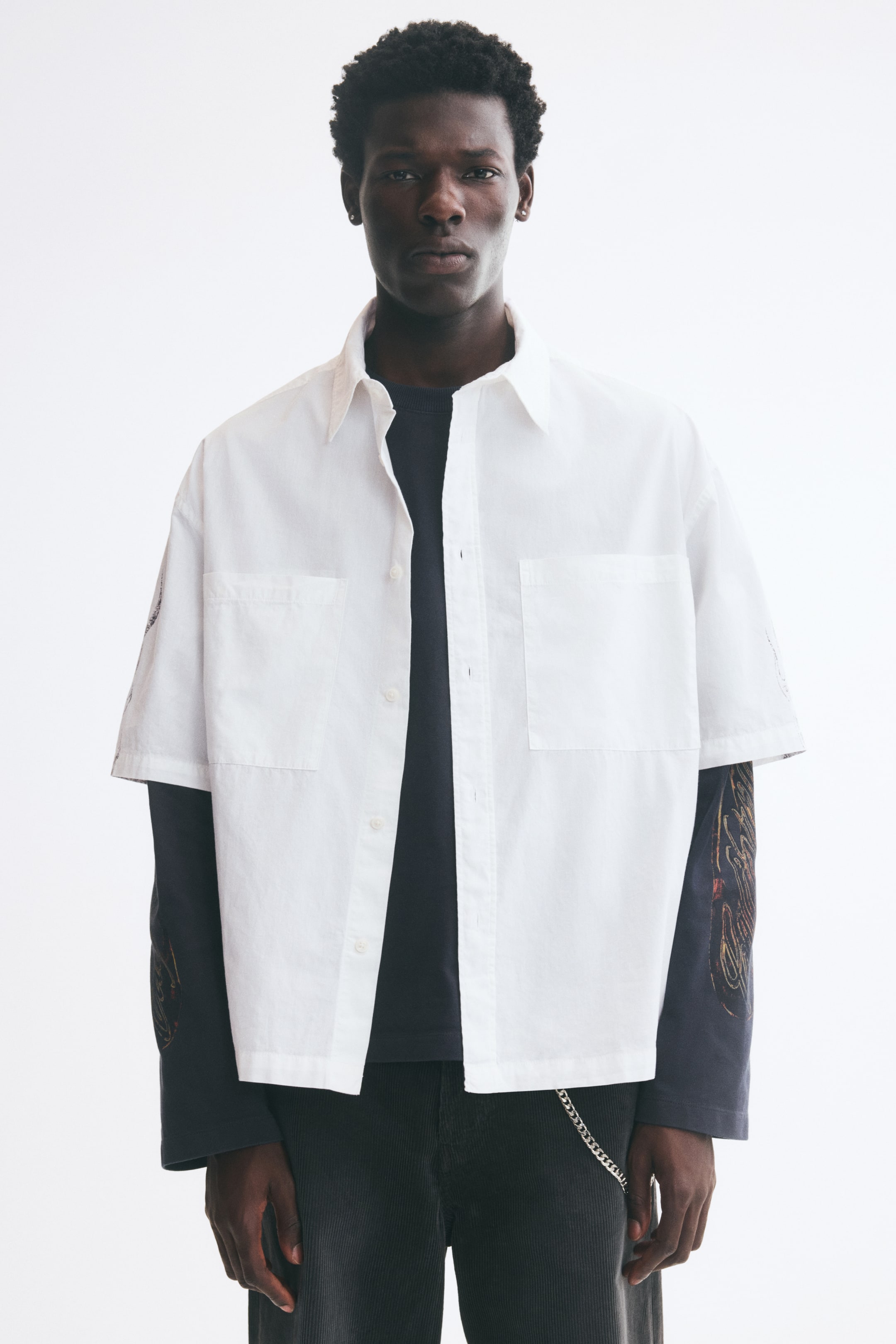 View larger image: A white short-sleeve cotton poplin shirt with subtle printed details on the sleeves is worn open by the model. The relaxed-fit shirt features a pointed collar, dropped shoulders, and chest patch pockets, layered over a dark long-sleeve top.