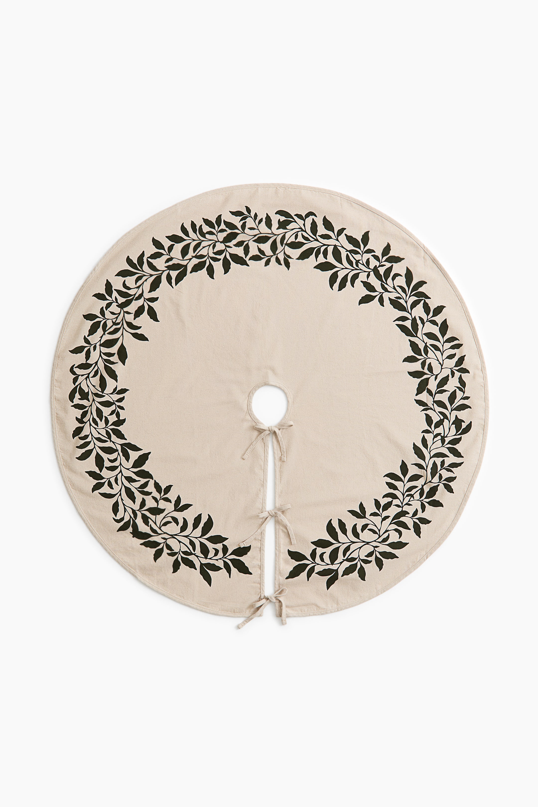View larger image: A circular beige tree skirt is shown from the front, featuring a dark green leafy vine pattern printed in a wreath design around the outer border. The skirt displays a central cutout and a straight opening secured by three fabric tie closures.