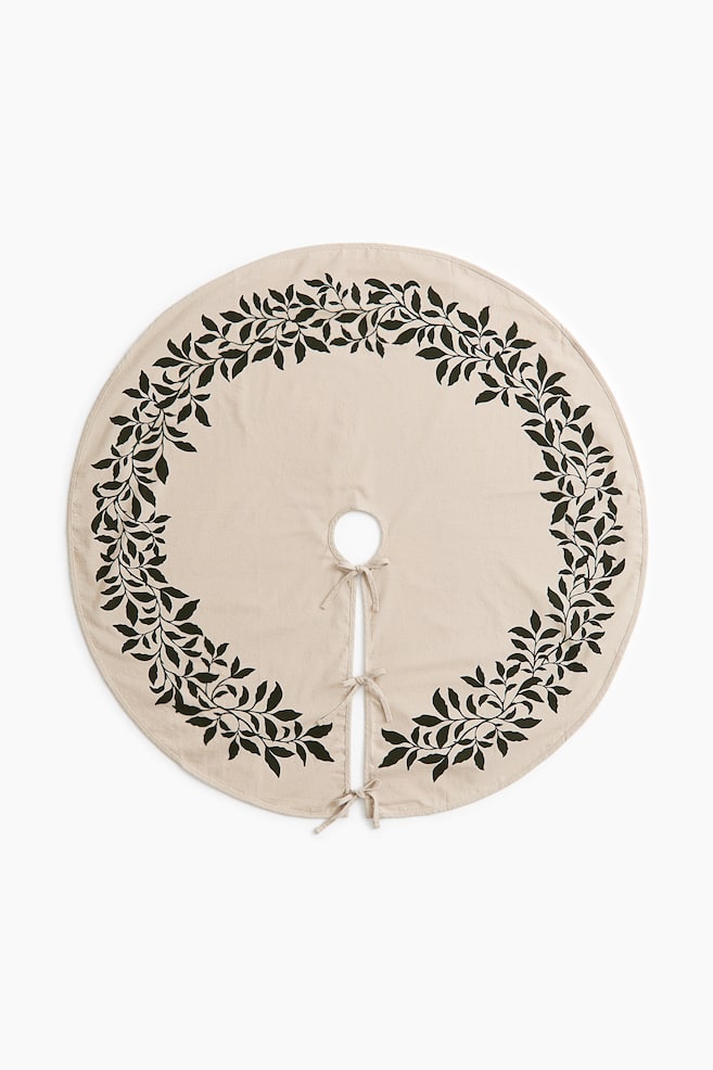A circular beige tree skirt is shown from the front, featuring a dark green leafy vine pattern printed in a wreath design around the outer border. The skirt displays a central cut-out and a straight opening secured by three fabric tie fastenings. - 1
