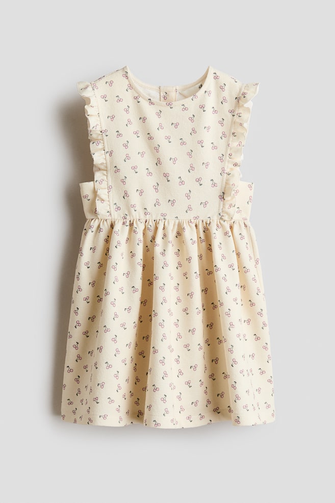 A sleeveless dress in textured cotton jersey is displaying its front, featuring a cream base with an all-over cherry print. Frill trims are detailing the front and extending over the shoulders, while a round neckline, gathered waist, and flared skirt are completing the design. - 1