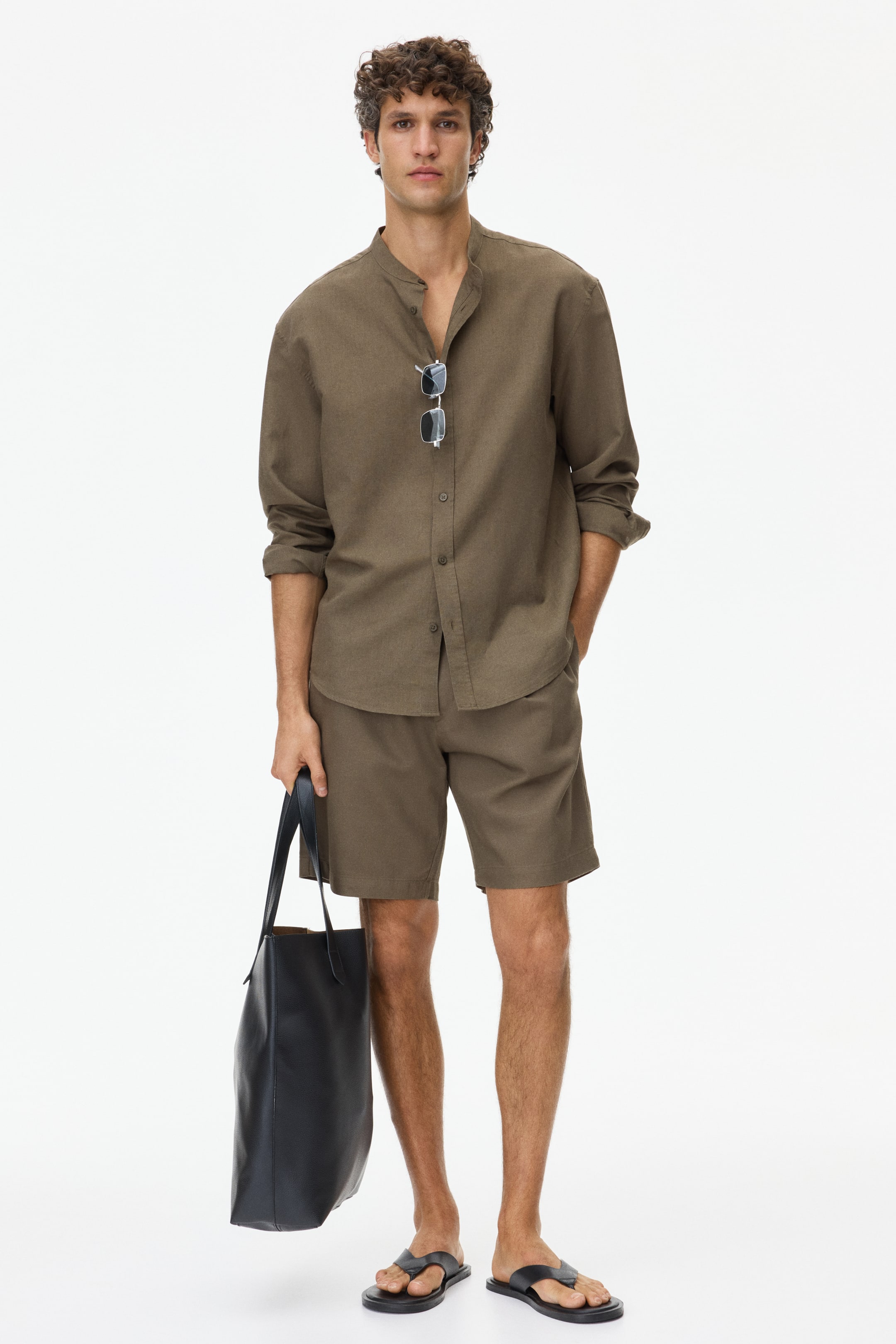 Agrandir l'image: Une chemise pour homme à col mao de couleur kaki brun et un short assorti sont présentés. La chemise à coupe décontractée est stylée avec des manches longues retroussées jusqu'aux avant-bras, et des lunettes de soleil pendent de sa patte de boutonnage. Le short assorti à coupe décontractée se termine au-dessus du genou.