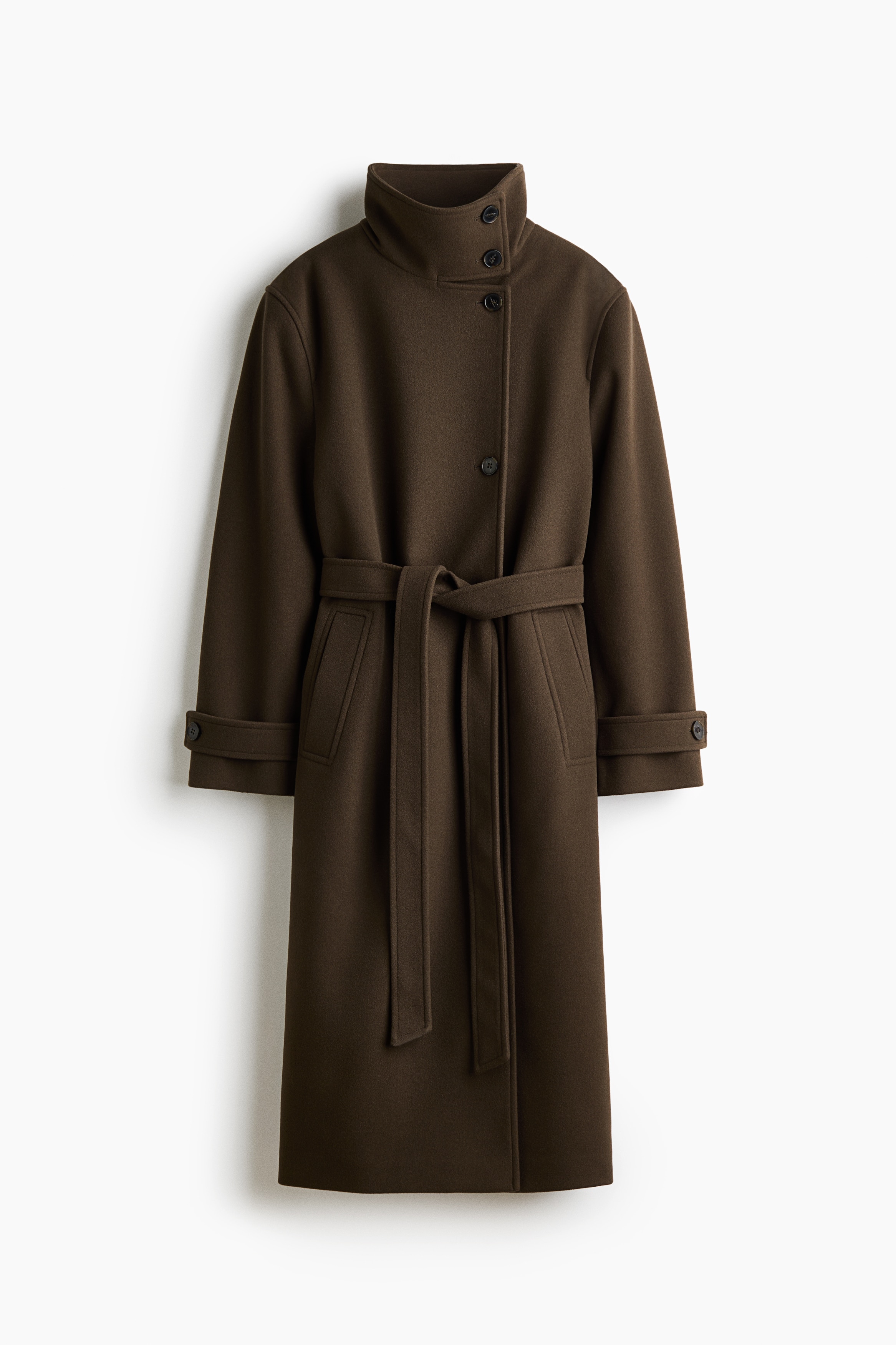 Het model draagt een donkerbruine, getextureerde oversized jas, met een hoge, gestructureerde opstaande kraag en een bijpassende ceintuur die in de taille is geknoopt. De jas heeft manchetten met knoopsluiting en is gestyled met het model dat één hand in een paspelzak aan de voorkant laat rusten.
