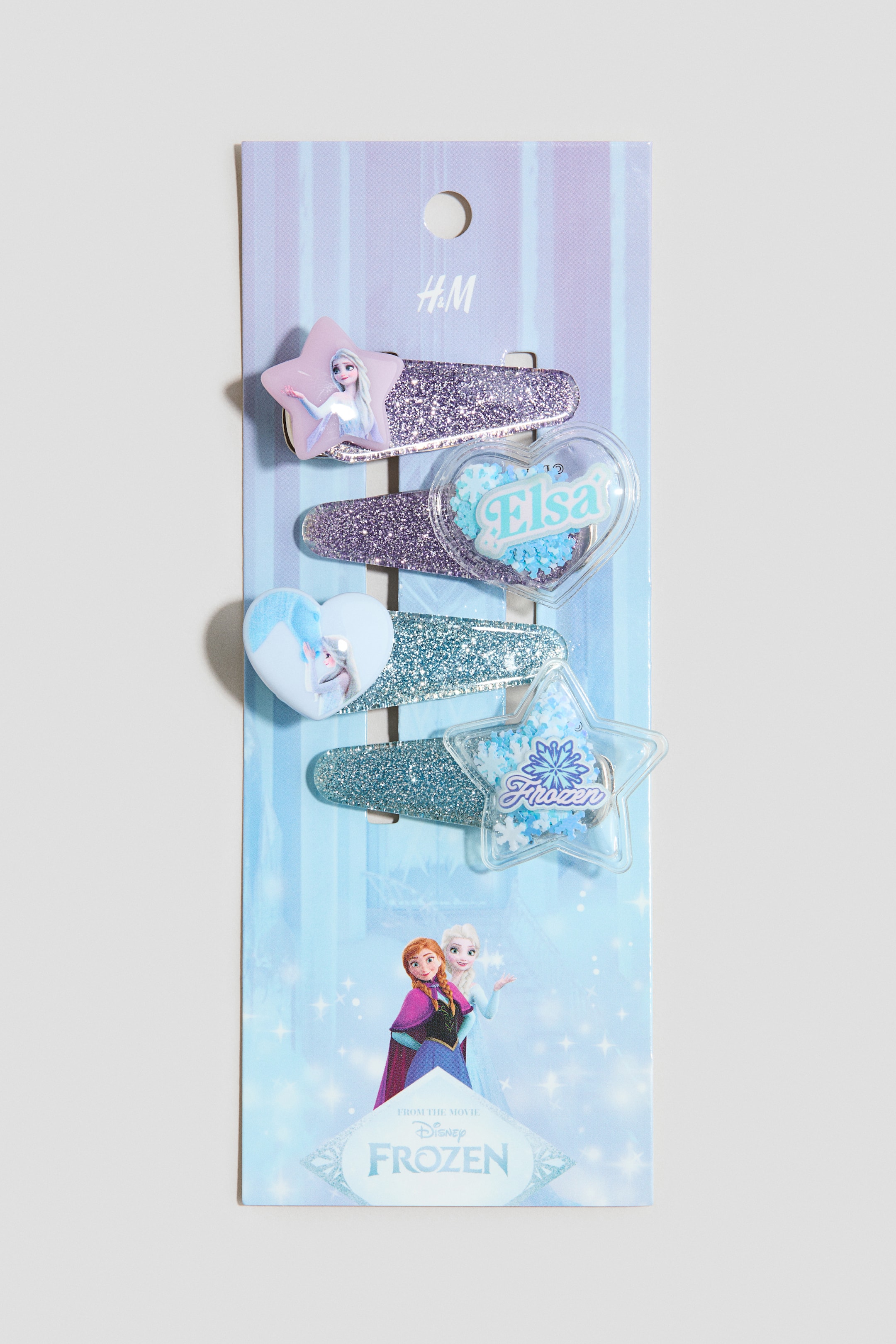 הצגת תמונה גדולה יותר: סט של ארבע סיכות שיער ממתכת מוצג מלפנים עם עיצובים שונים. שתי הסיכות מעוטרות בנצנצים סגולים: אחת עם כוכב ורוד המציג את הדמות Elsa, והשנייה עם לב שקוף שמכיל את הטקסט "Elsa" ונצנצים כחולים. שתי הסיכות הנותרות מעוטרות בנצנצים תכולים: אחת עם לב תכול המציג את הדמות Anna, והשנייה עם כוכב שקוף שמכיל את הטקסט "Frozen" ונצנצים כחולים.