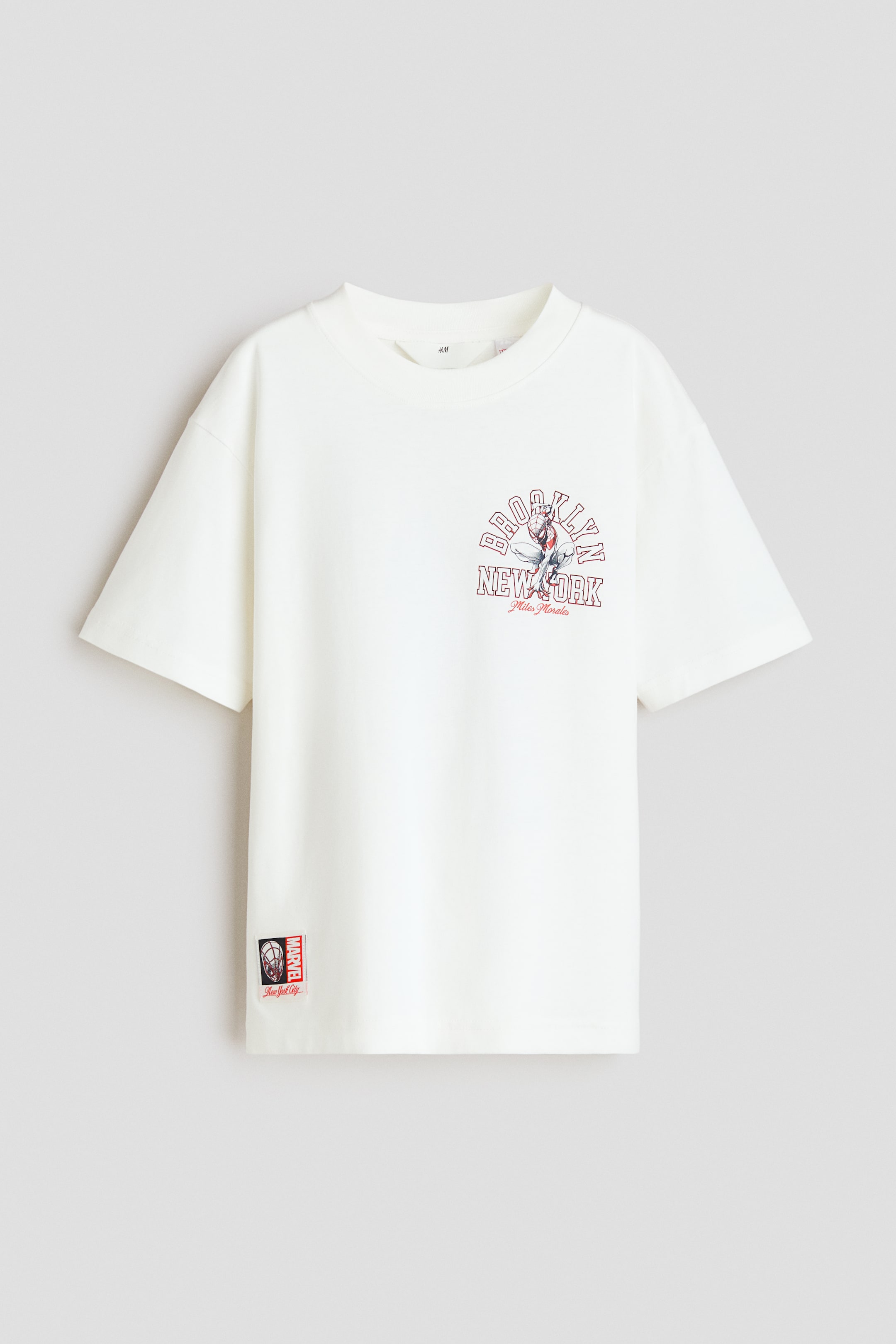 拡大画像を表示: クリームカラーのコットンジャージーTシャツは、ネックライン周りにリブトリムを施し、ゆったりとしたフィット感とドロップショルダーが特徴です。中央にはスパイダーマン(マイルズ・モラレス)が描かれた「ブルックリン ニューヨーク」のグラフィックがあり、左下には小さなマーベルのタグが見えます。