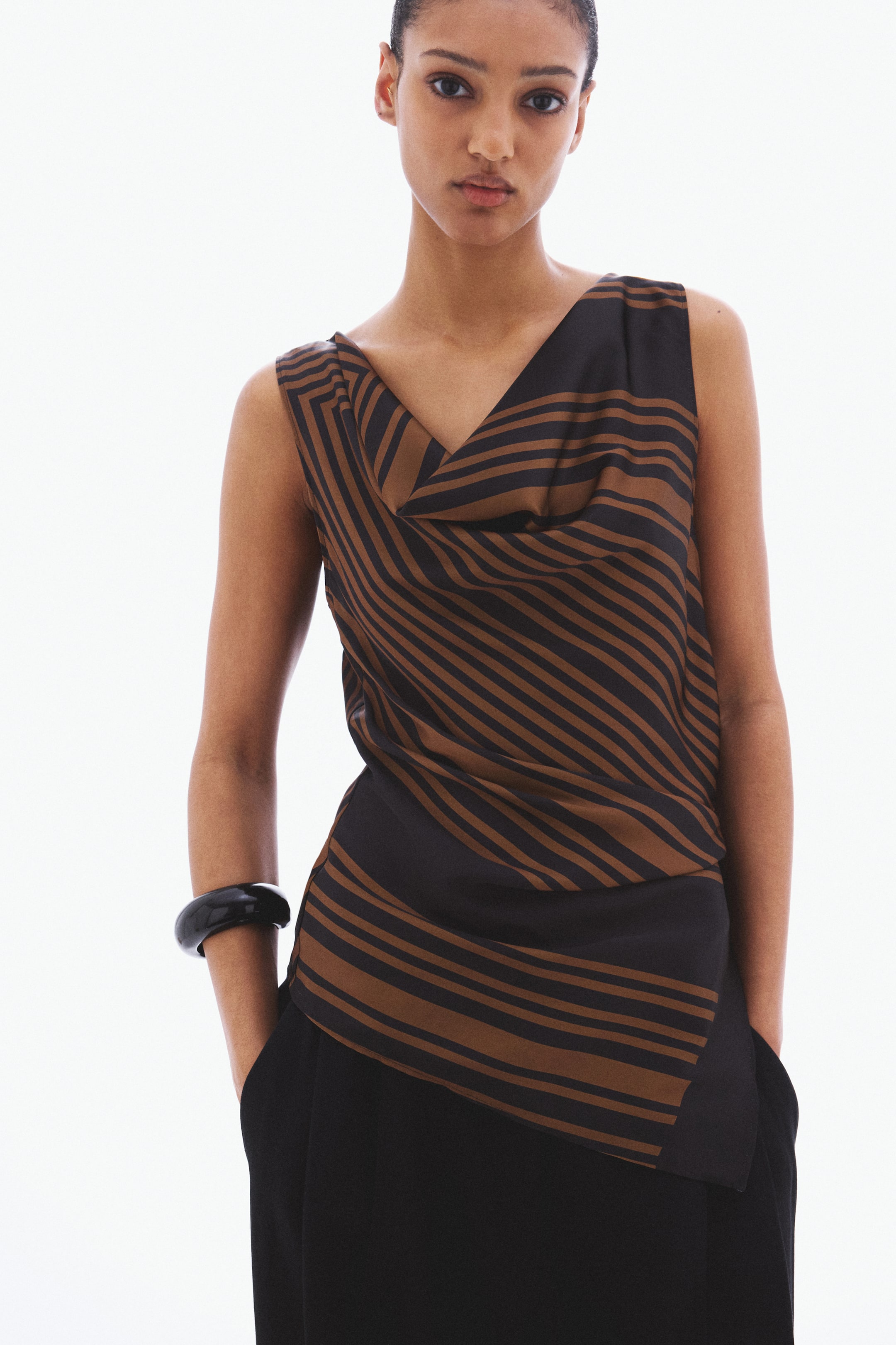 View larger image: A sleeveless top is facing forward and features a black and brown angled striped pattern. It showcases a draped cowl neckline and an asymmetrical hem, layered over black trousers.