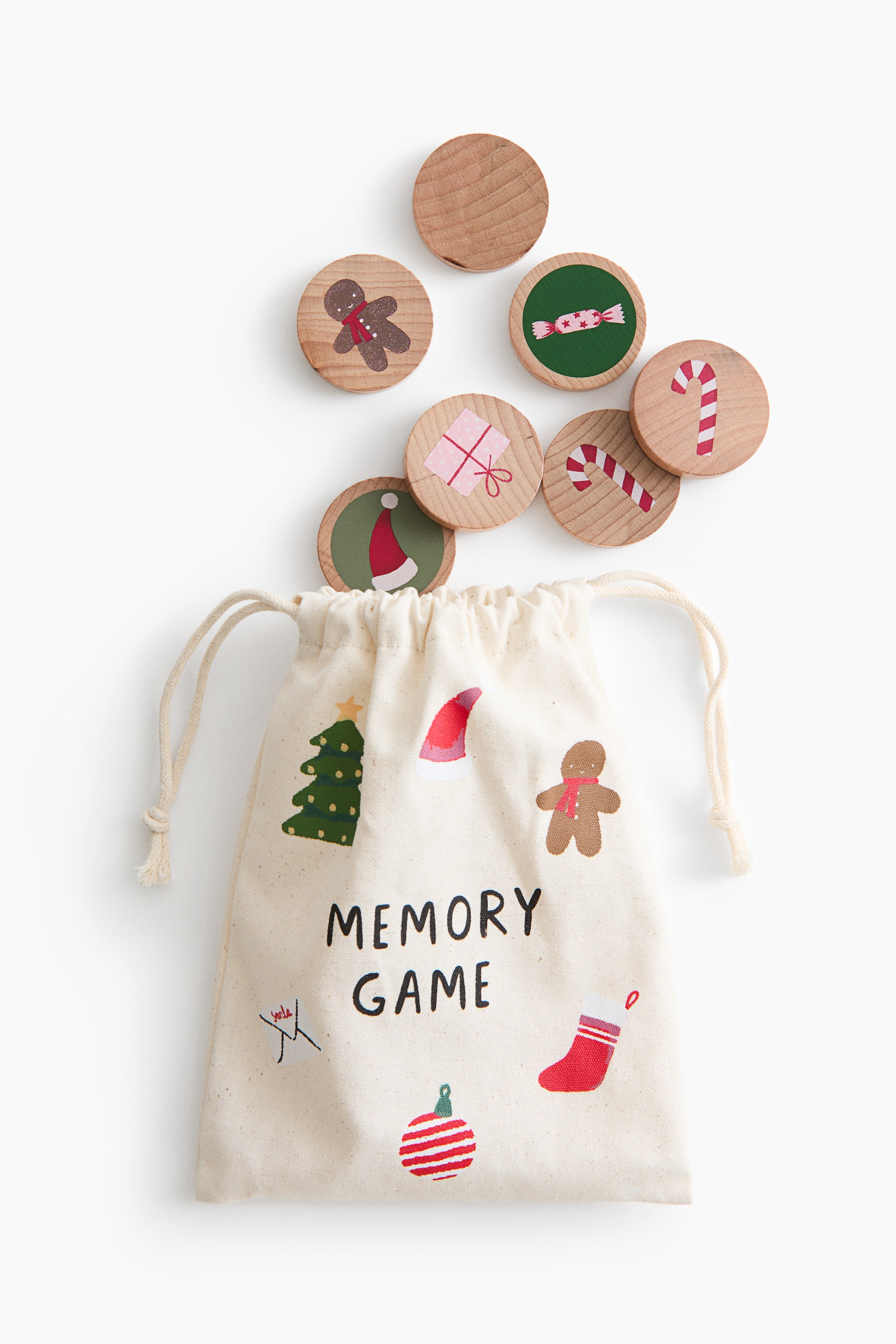 View larger image: An off-white fabric drawstring bag featuring festive illustrations and the words "MEMORY GAME" lies flat, with several round wooden game tokens appearing to spill out from its top opening. The wooden tokens display various holiday-themed prints, including a gingerbread man, candy canes, a Santa hat, and a wrapped gift.