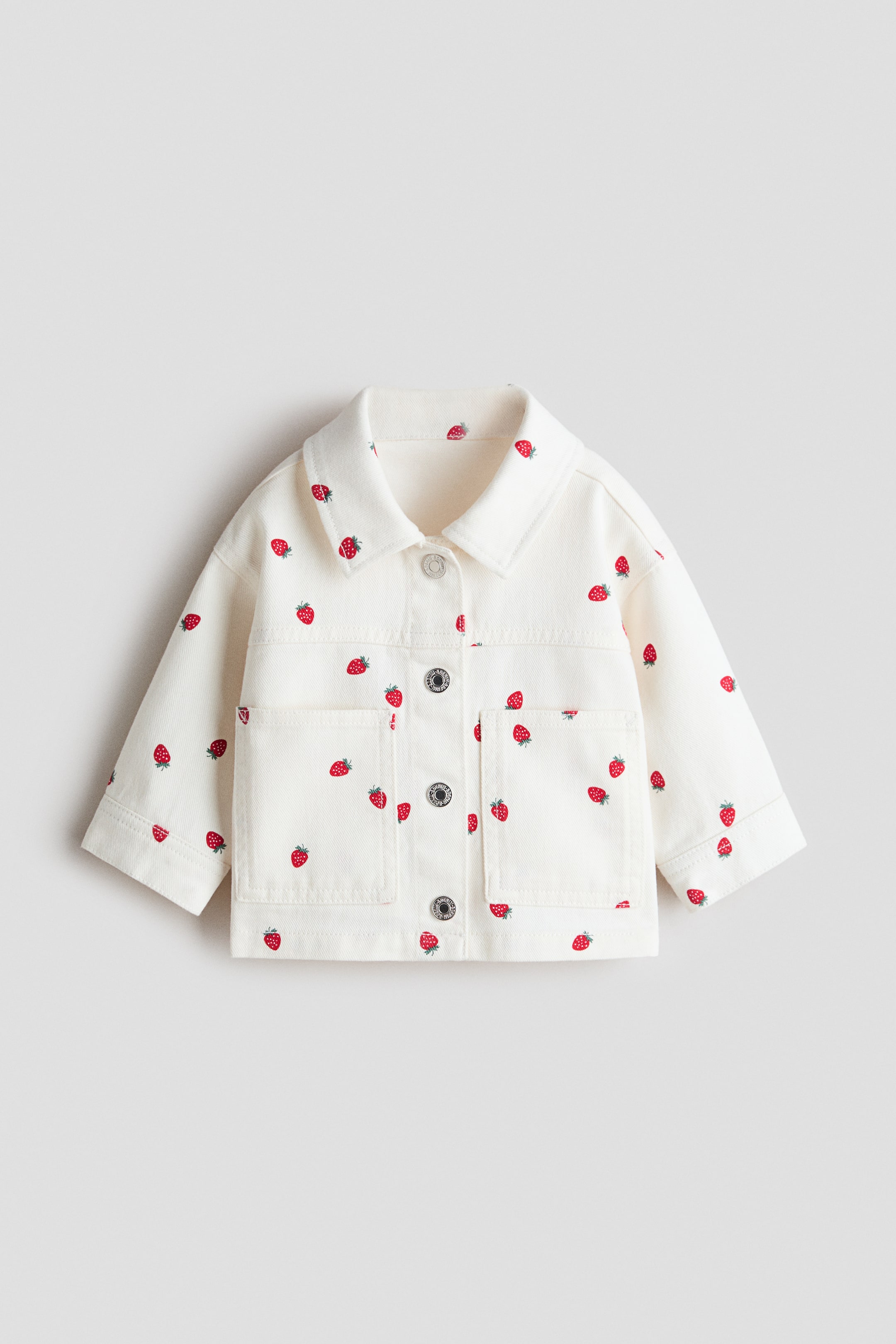 Grotere afbeelding bekijken: Een off-white jas is frontaal afgebeeld (FRONT) en heeft een all-overprint van rode aardbeien met groene steeltjes. Hij heeft een klassieke kraag, een drukknoopsluiting aan de voorzijde met zilverkleurige knopen en vier opgestikte zakken.