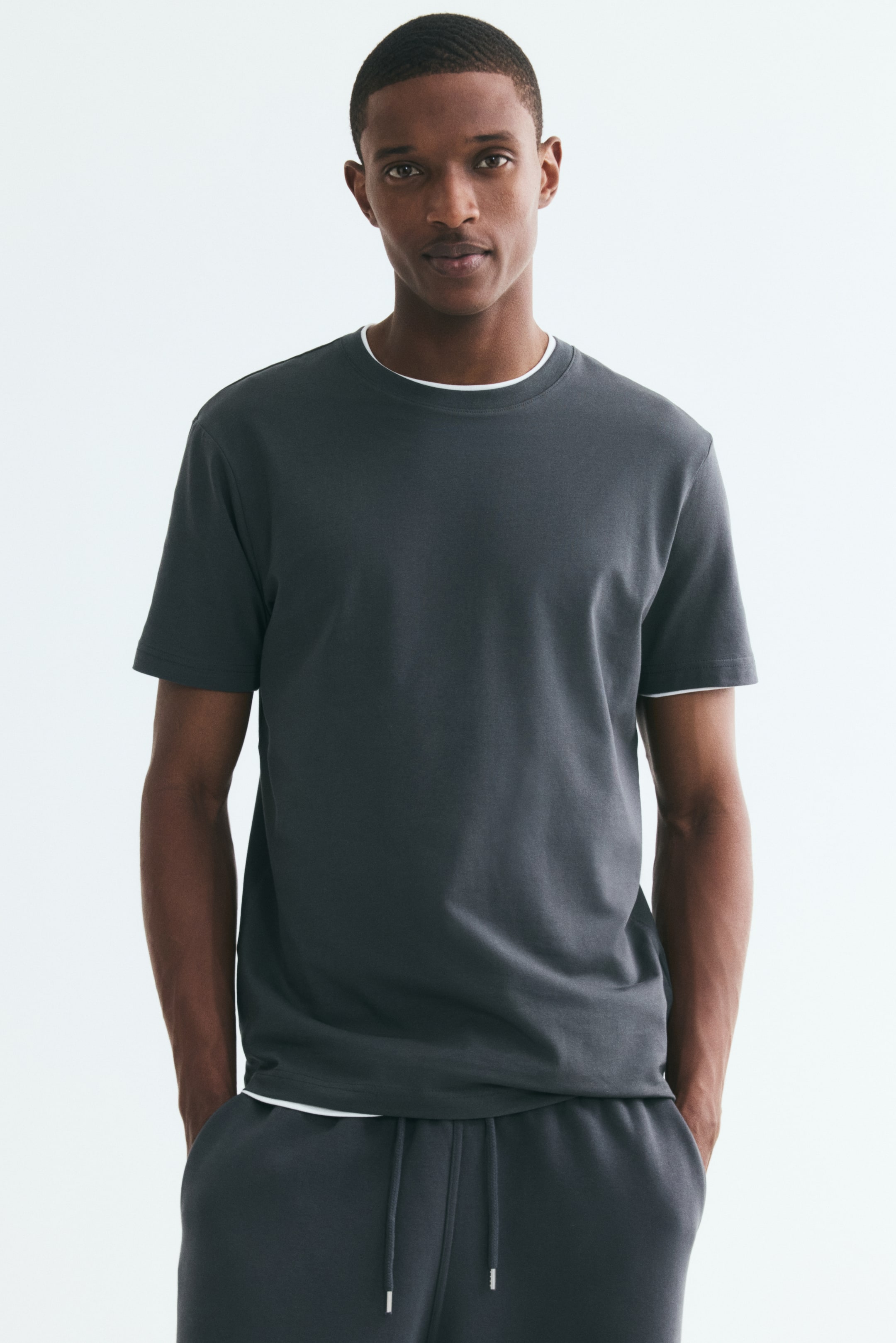 View larger image: The model is wearing a dark grey slim-fit T-shirt featuring a round, rib-trimmed neckline and a straight hem. The T-shirt is layered over a white undershirt, visible at the collar and sleeve cuffs, and is paired with matching dark grey drawstring sweatpants.