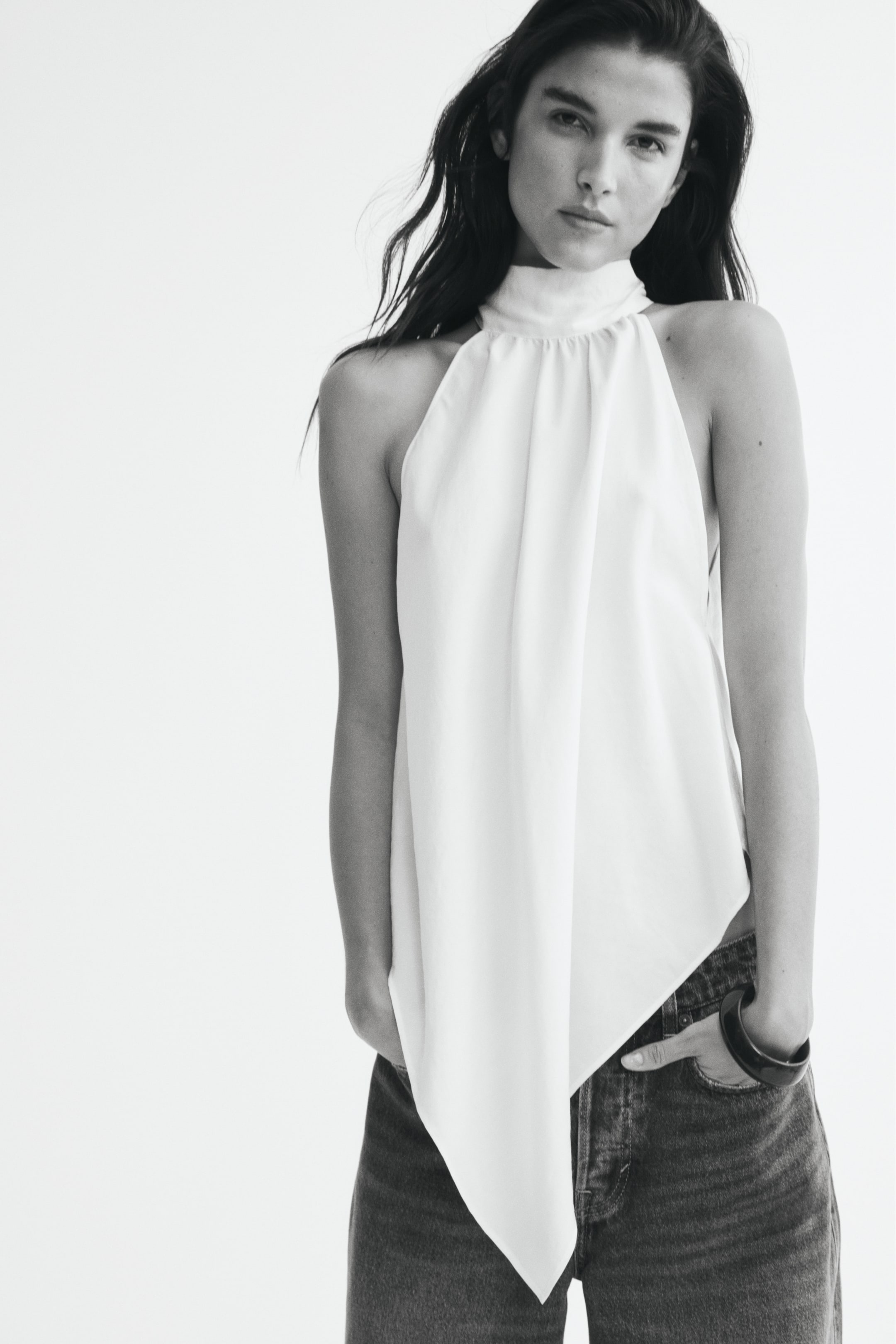 Προβολή μεγαλύτερης εικόνας: Ένα αμάνικο halter top από ελαφρύ υφαντό ύφασμα φοριέται από το μοντέλο, με μαζεμένο όρθιο γιακά και ντραπέ μπροστινό μέρος που καταλήγει σε μακρύ, μυτερό τελείωμα σε σχήμα V. Το top συνδυάζεται με σκούρο denim τζιν, ενώ το μοντέλο στέκεται με τα χέρια τοποθετημένα στις μπροστινές τσέπες.