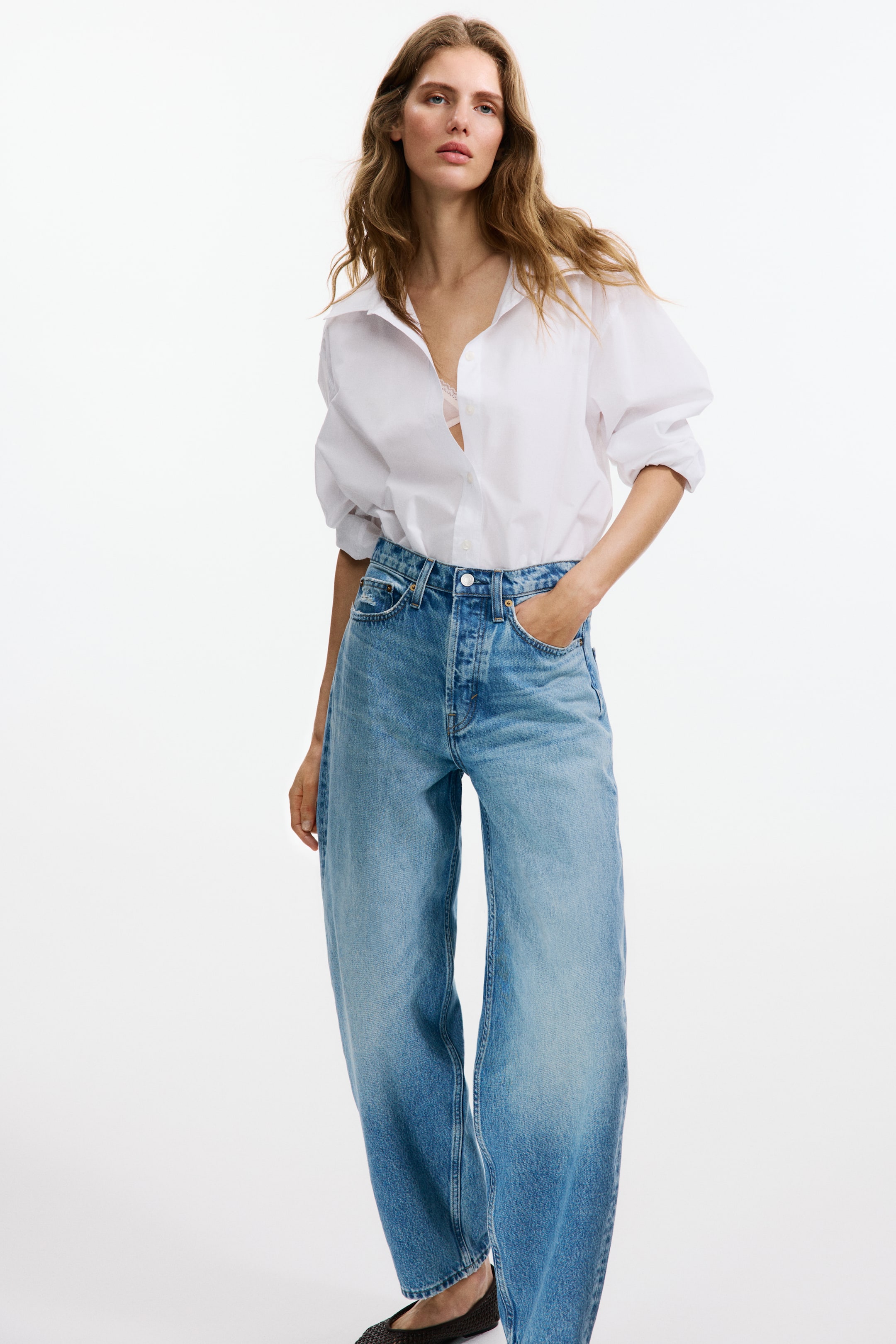 View larger image: A white button-down shirt featuring a spread collar, open at the neckline, subtly revealing a light-coloured bra, and rolled-up voluminous sleeves. It is worn with light wash blue wide-leg jeans, showcasing a button fly fastening and a relaxed fit through the leg.