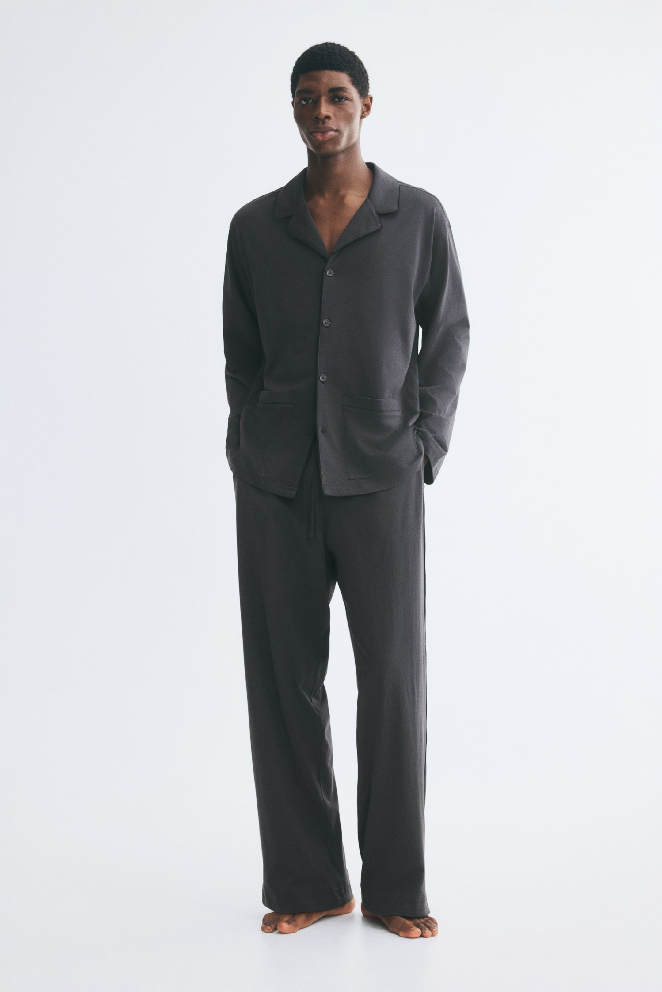 View larger image: The model is wearing a dark grey cotton jersey pyjama set. The set features a long-sleeved, button-down shirt with a resort collar and front patch pockets, paired with matching loose-fit trousers with a drawstring waist and side pockets.