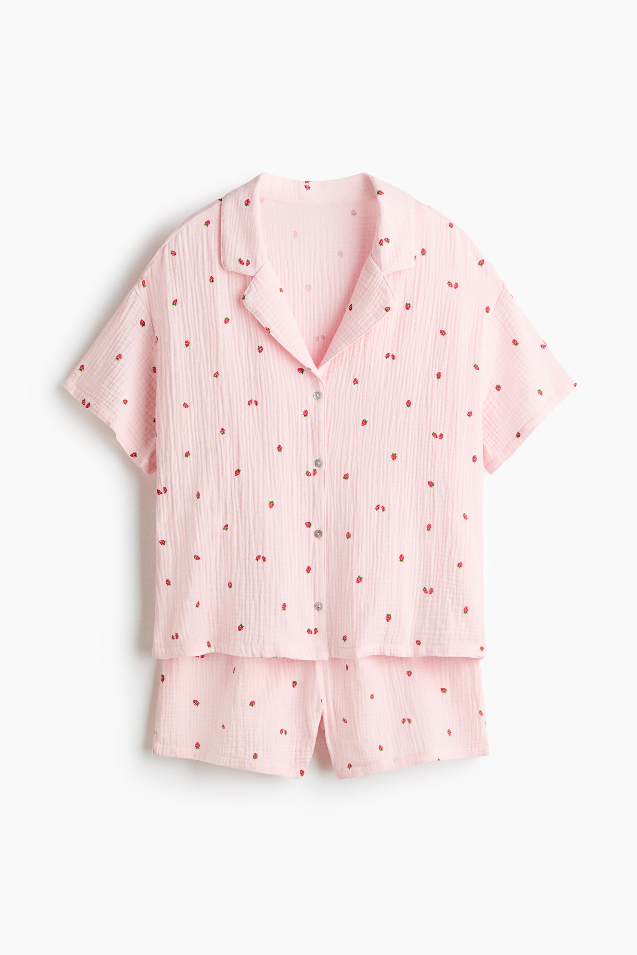 Ver imagen más grande: Un conjunto de pijama rosa se muestra en vista frontal, con una camiseta de manga corta y pantalones cortos a juego, ambos adornados con un pequeño estampado de fresas rojas. La camiseta presenta un cuello de solapa y un cierre frontal de botones, mientras que ambas piezas exhiben una tela con textura arrugada.