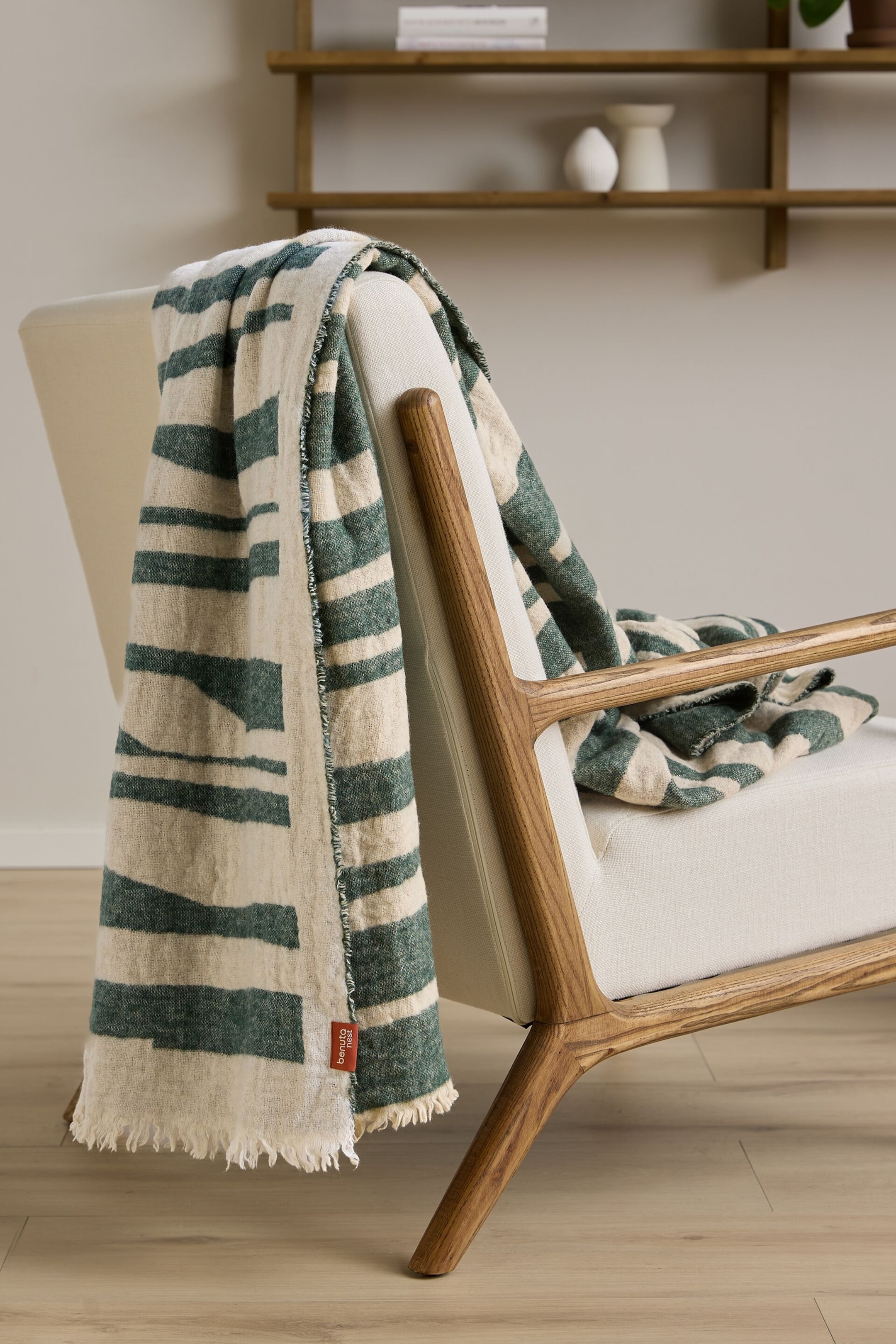 Agrandir l'image: Une couverture crème et verte est drapée sur un fauteuil, présentant des rayures horizontales irrégulières, des bords effilochés et une étiquette rouge « Benuta next » visible sur un bord.
