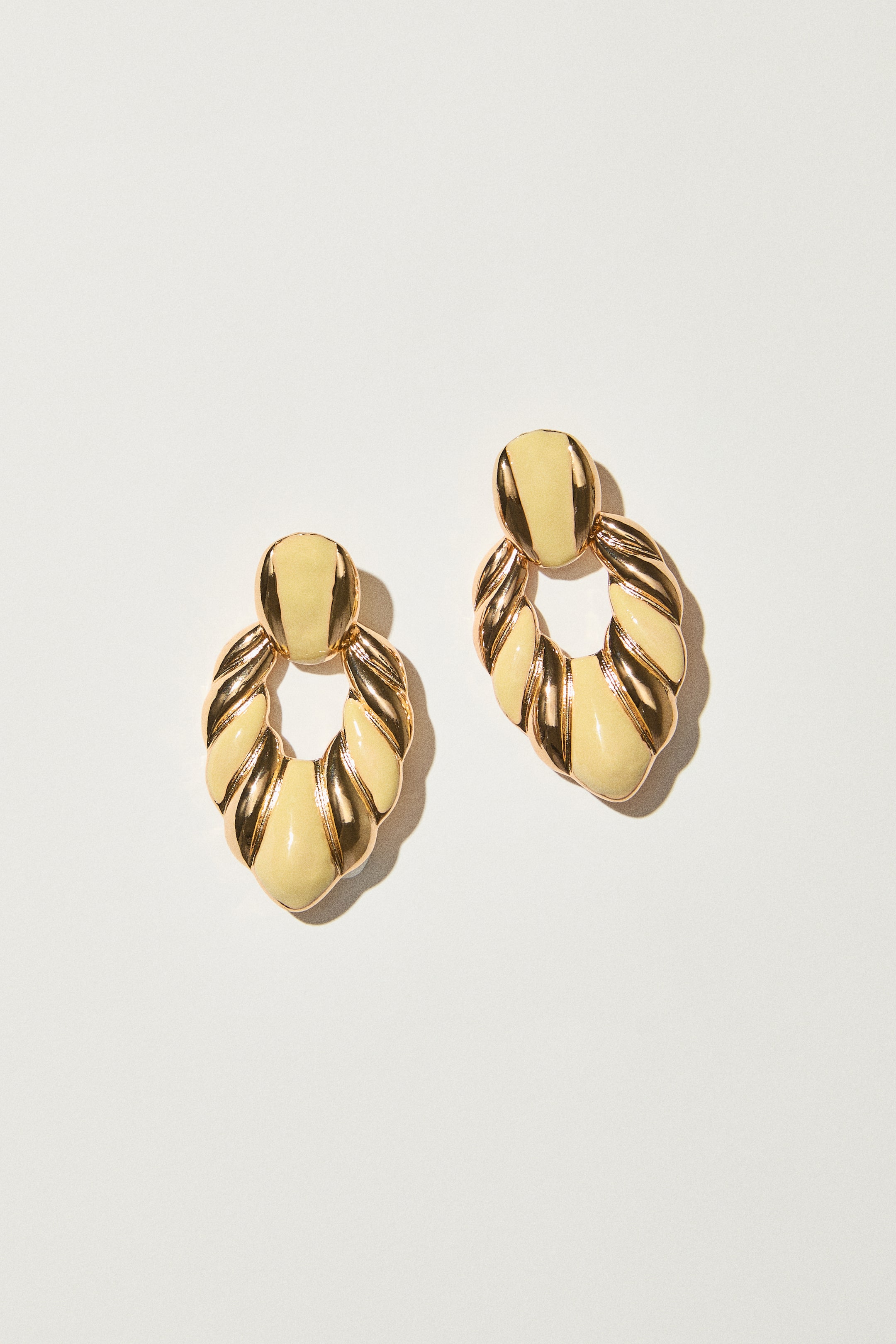 Agrandir l'image: Une paire de boucles d'oreilles en métal doré et émail jaune pâle. Chaque boucle d'oreille présente un clou ovale relié à un pendentif ovale plus grand, torsadé et creux, mettant en valeur des sections alternées de métal doré brillant et d'émail lisse.