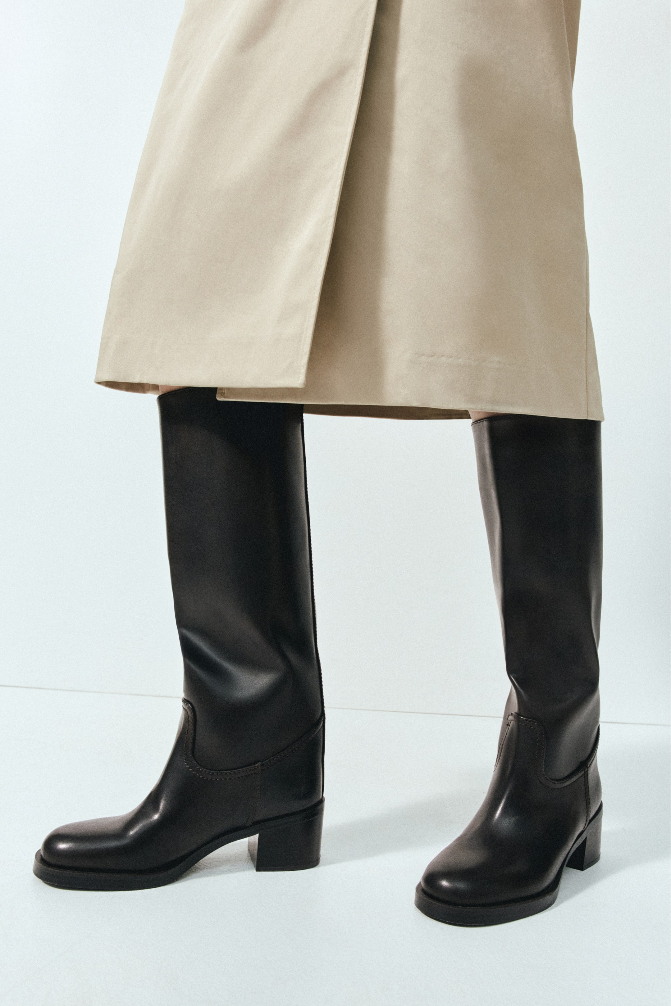 View larger image: The model is wearing knee-high black leather boots with rounded toes and low block heels. The boots are styled with a beige wrap skirt that falls just above the knee.