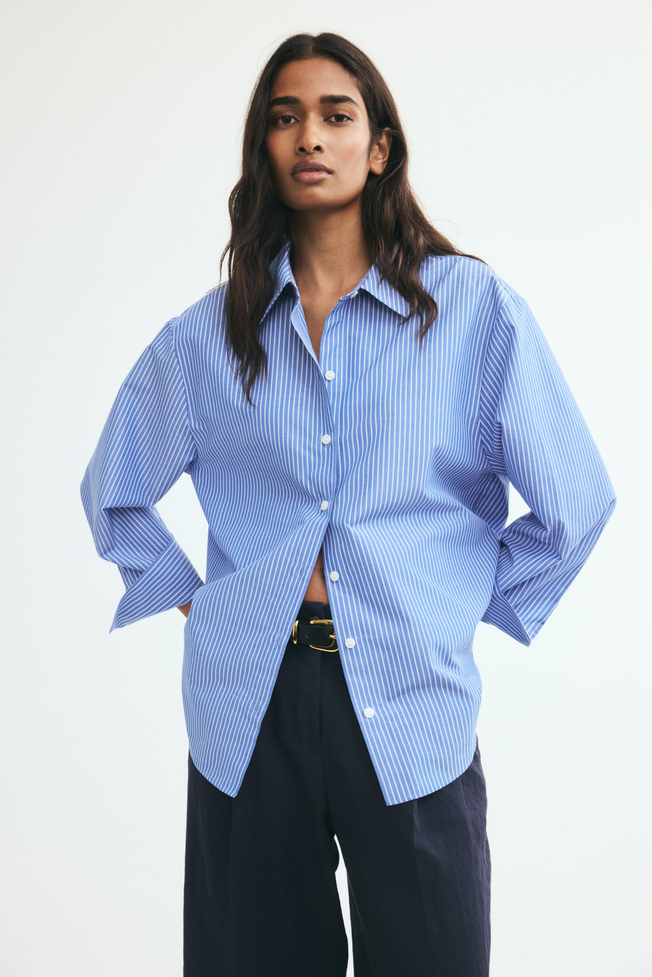 View larger image: A blue and white vertically striped woven shirt featuring a collar, dropped shoulders, and long sleeves is worn by the model. The shirt is partially unbuttoned at the lower front, showcasing its gently rounded hem draped over dark trousers with a belted waist.