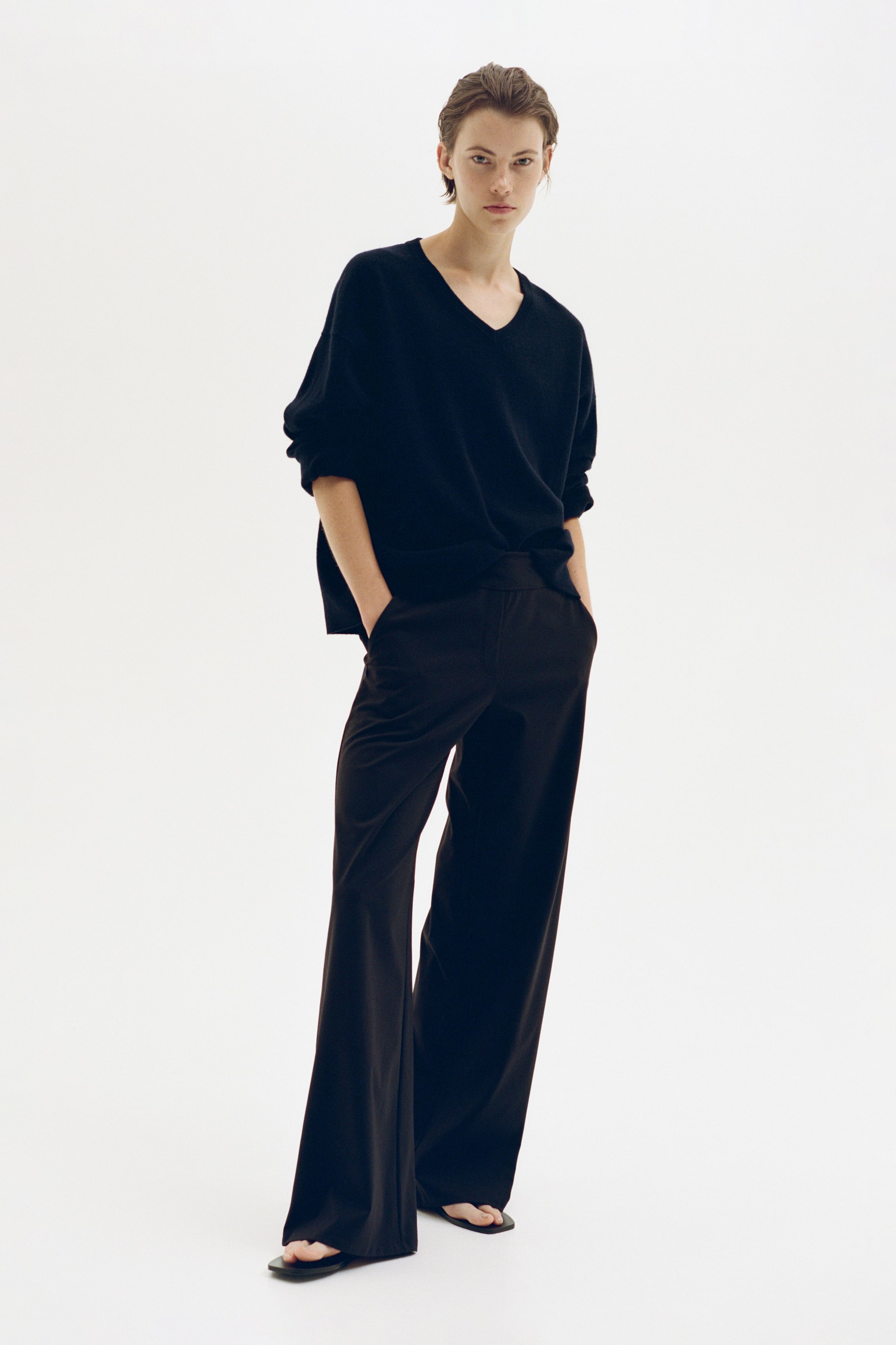 View larger image: Wide-leg pants in a dark brown shade are shown from the front, featuring a relaxed fit with a fluid drape, an elasticised waistband, and a mock fly. The pants are styled with a black V-neck sweater, highlighting their soft texture and comfortable fit.