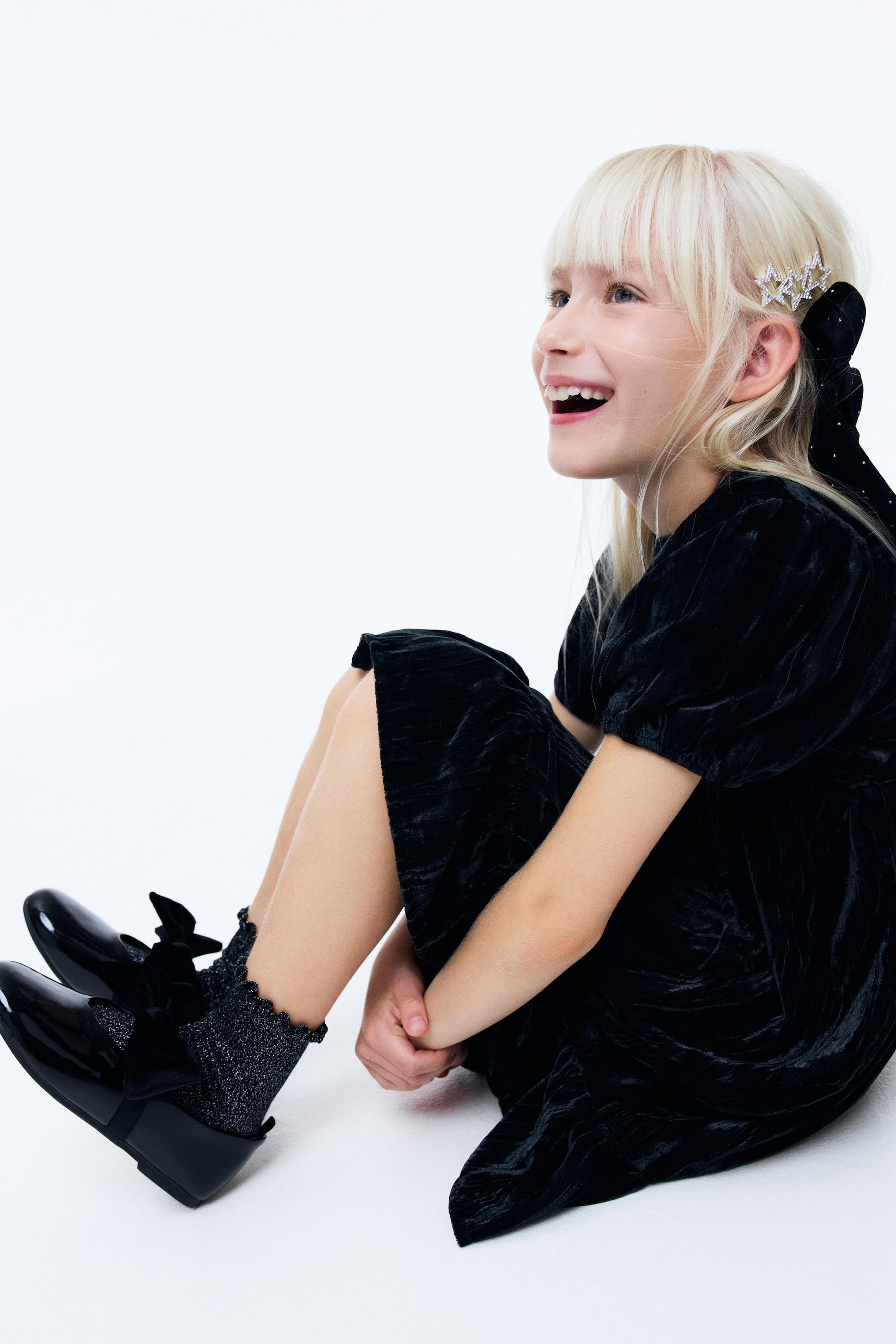 View larger image: A black velvet dress is featuring short puffed sleeves. The dress is draping loosely, and its skirt is gathering around the legs, showcasing its soft texture and subtle sheen from a side-front angle.