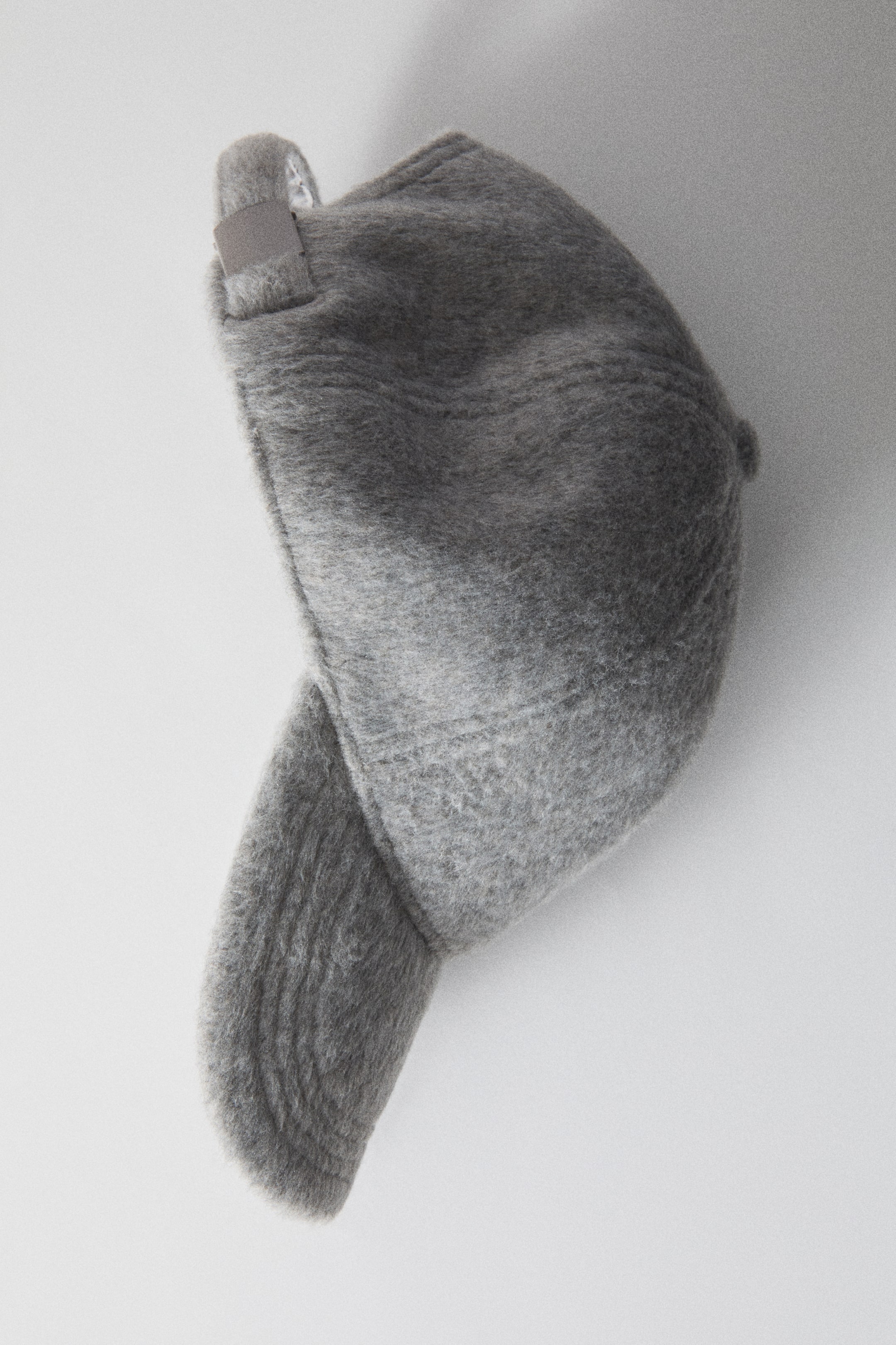 Ampliar la imagen: Se muestra una gorra de tela afieltrada gris en vista de perfil lateral, destacando su superficie con textura esponjosa y su visera curva. La parte trasera de la gorra cuenta con una correa ajustable con un broche de metal.
