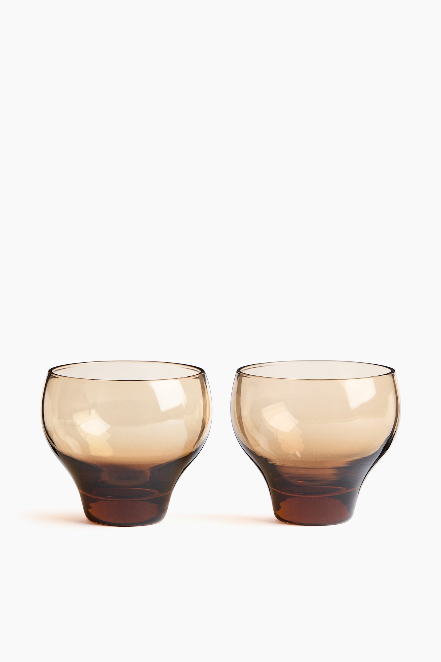 A pair of mouth-blown glass tumblers in a translucent amber-brown hue is standing side-by-side from a front angle, showcasing rounded bowls tapering down to thick, solid bases. The glass is displaying a gradient color effect that deepens toward the bottom of each tumbler.