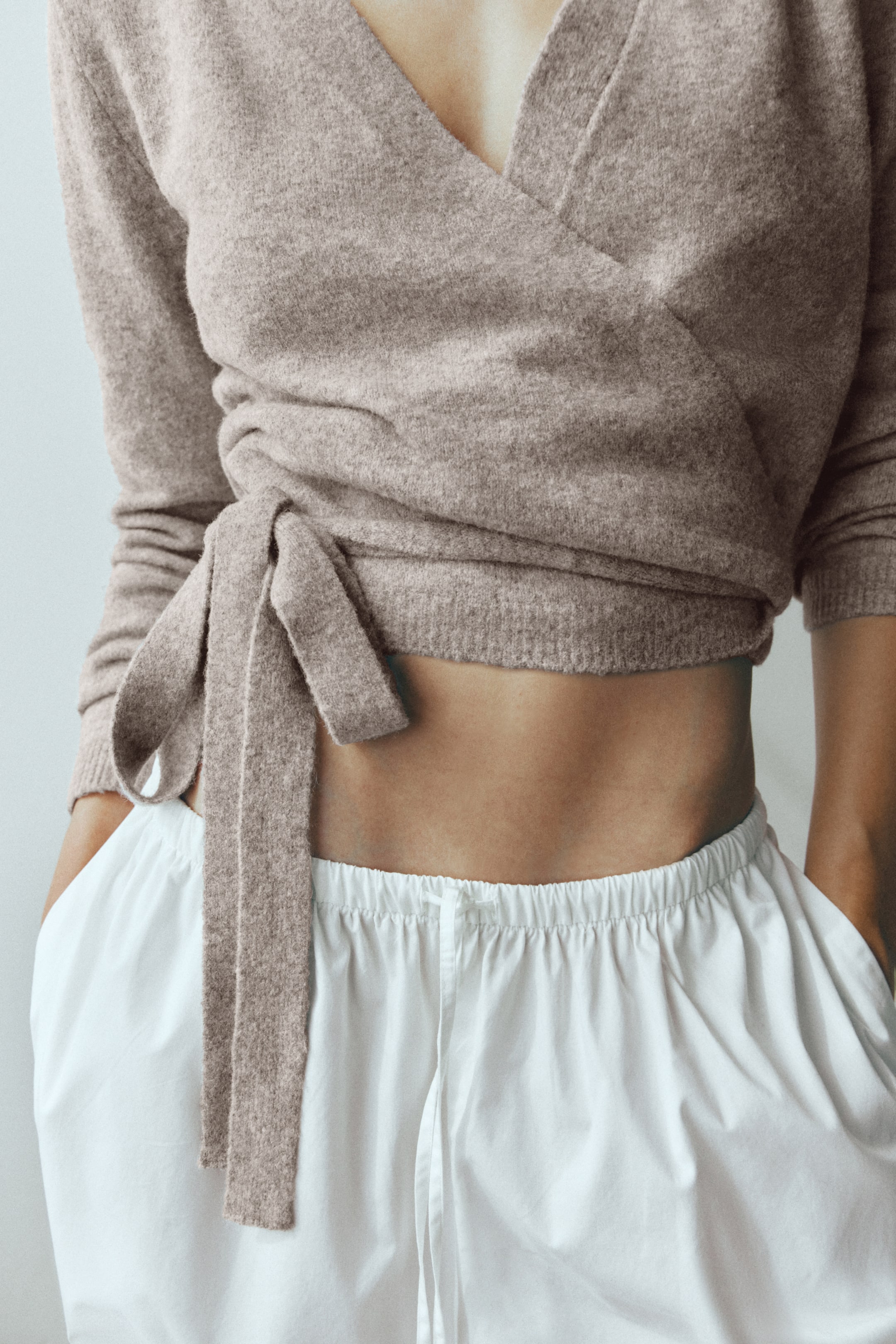 Se større billede: En kort, lysebrun strik-wrap-cardigan bæres af modellen og har V-udskæring, lange ærmer og brede bindebånd i stof, der er fastgjort i den ene side. Cardiganen er stylet med hvide bukser med snøre i taljen, mens modellen har hænderne i lommerne.
