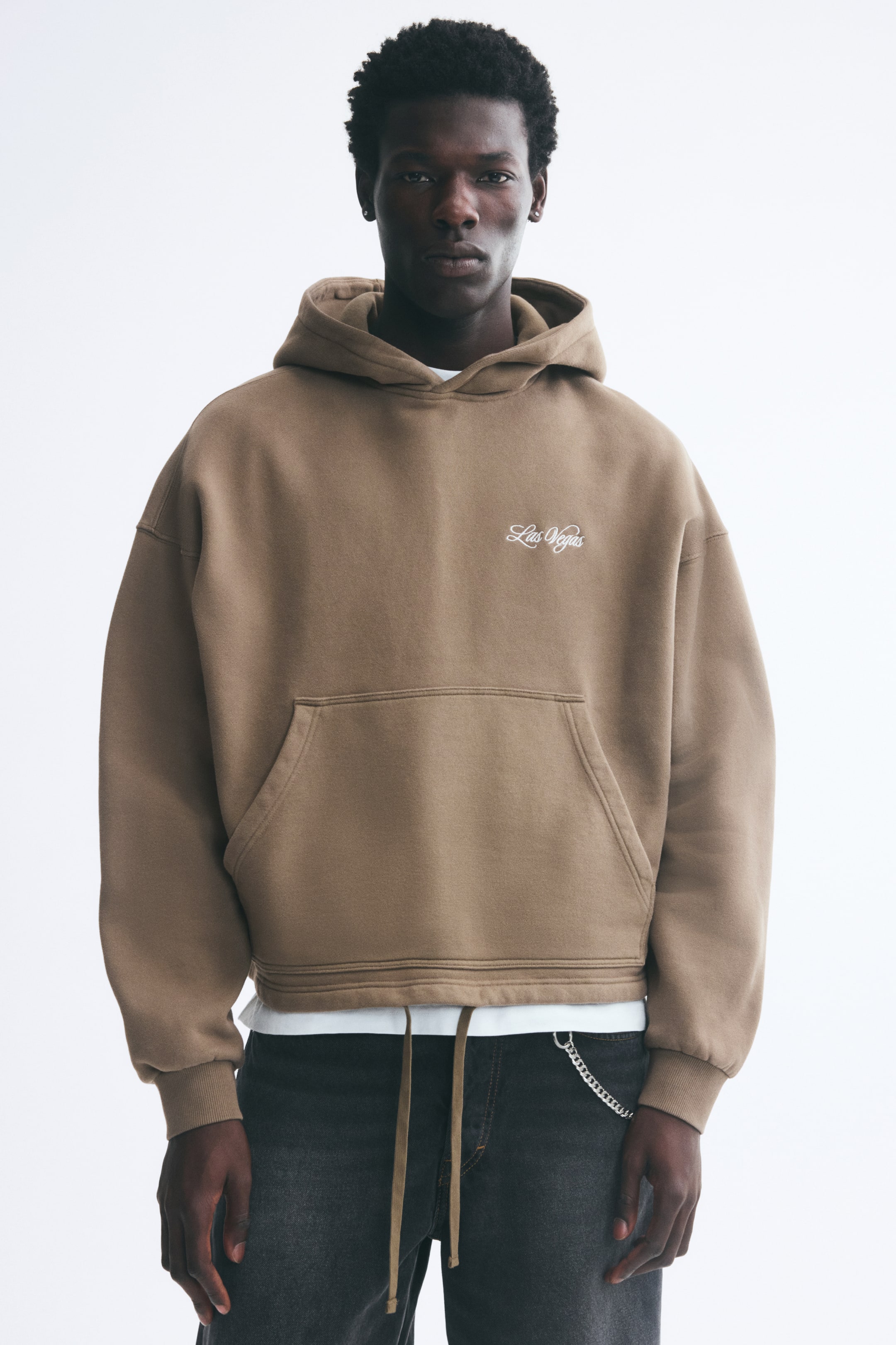 View larger image: The model is wearing a brown oversized hoodie featuring a wrap hood, dropped shoulders, a kangaroo pocket, and a small white script detail on the chest. The hoodie is layered over a white T-shirt and paired with dark jeans, with drawstrings hanging from the hem.