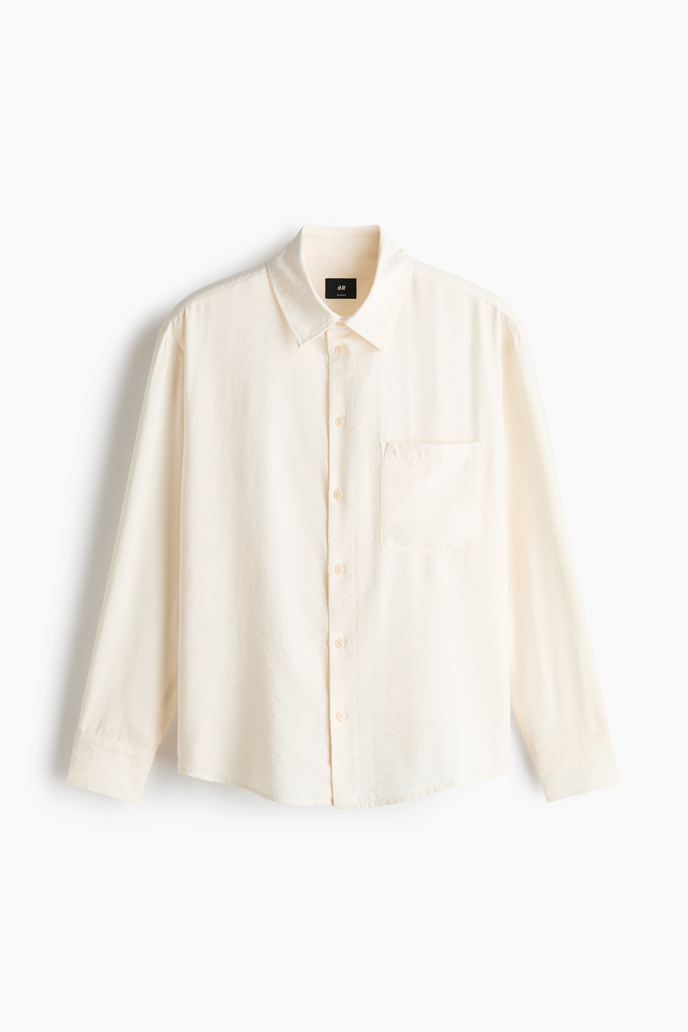 The model is wearing a light cream-coloured shirt in a crinkled weave, featuring a relaxed fit, a collar, a buttoned front, and a single patch chest pocket. The long-sleeved shirt is styled untucked with matching light-coloured trousers and a patterned neck scarf.