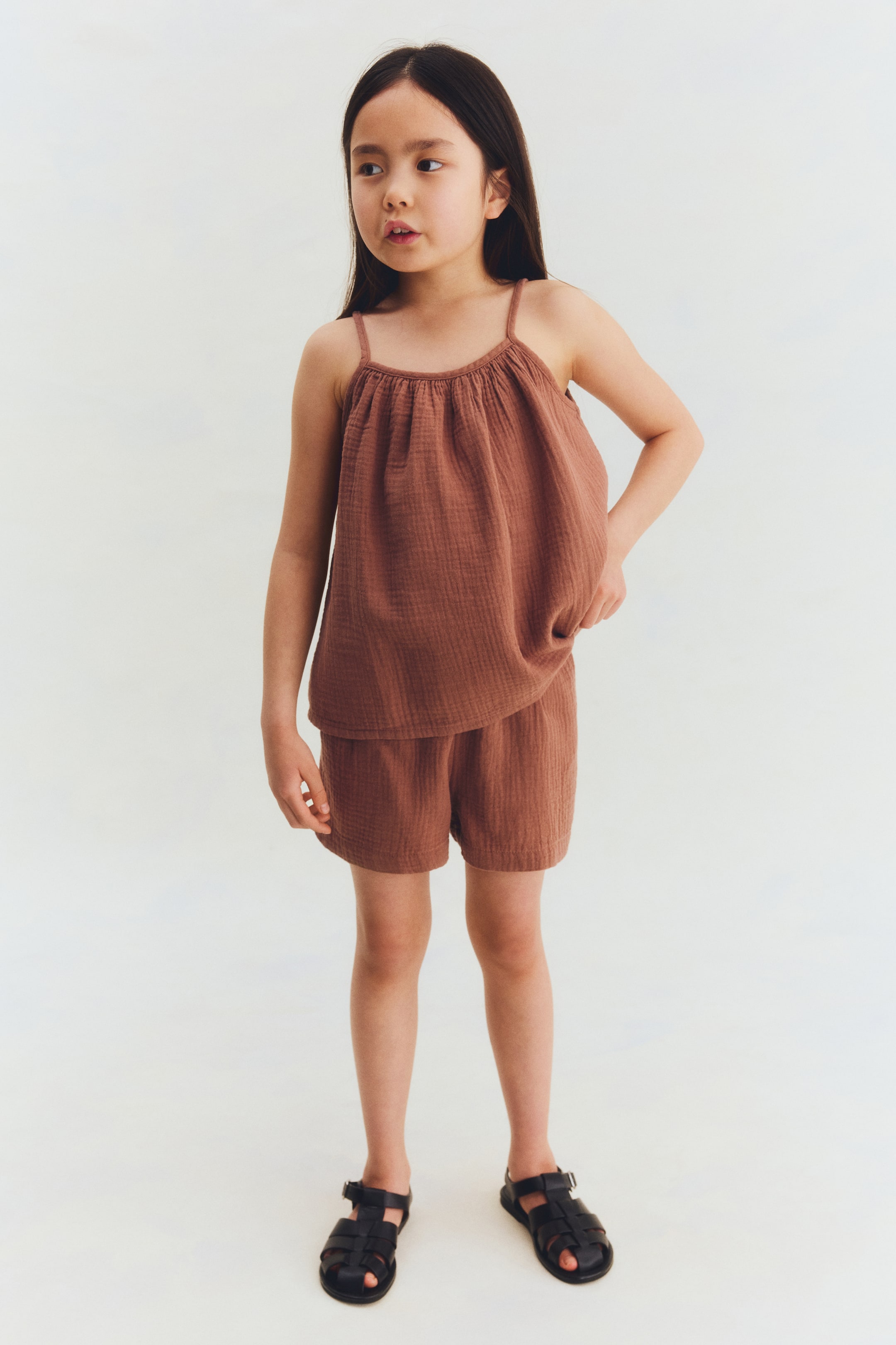 Visualizza immagine più grande: Un completo per bambini a due pezzi di colore ruggine viene mostrato in vista frontale, composto da un top canotta con spalline sottili e scollo arricciato, e pantaloncini abbinati dal taglio ampio. Il tessuto presenta una texture increspata e garzata.