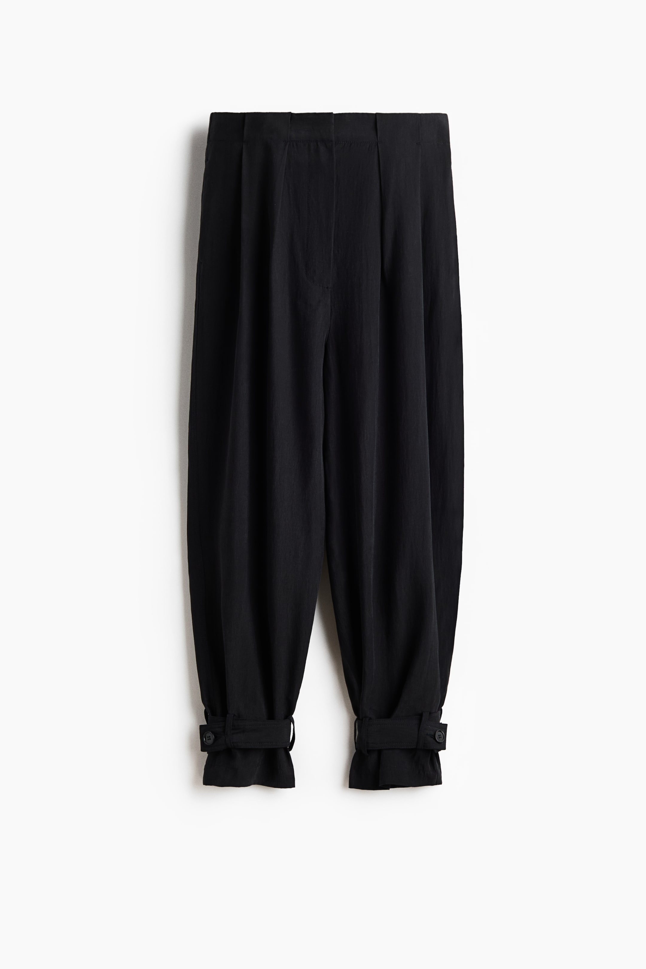 Ver imagen más grande: Pantalones negros de corte holgado en tejido de punto, presentados de frente en posición plana. Cuentan con cintura alta, bragueta con cremallera, piernas tipo globo con pliegues en la parte superior y lengüetas ajustables con botones en los dobladillos.