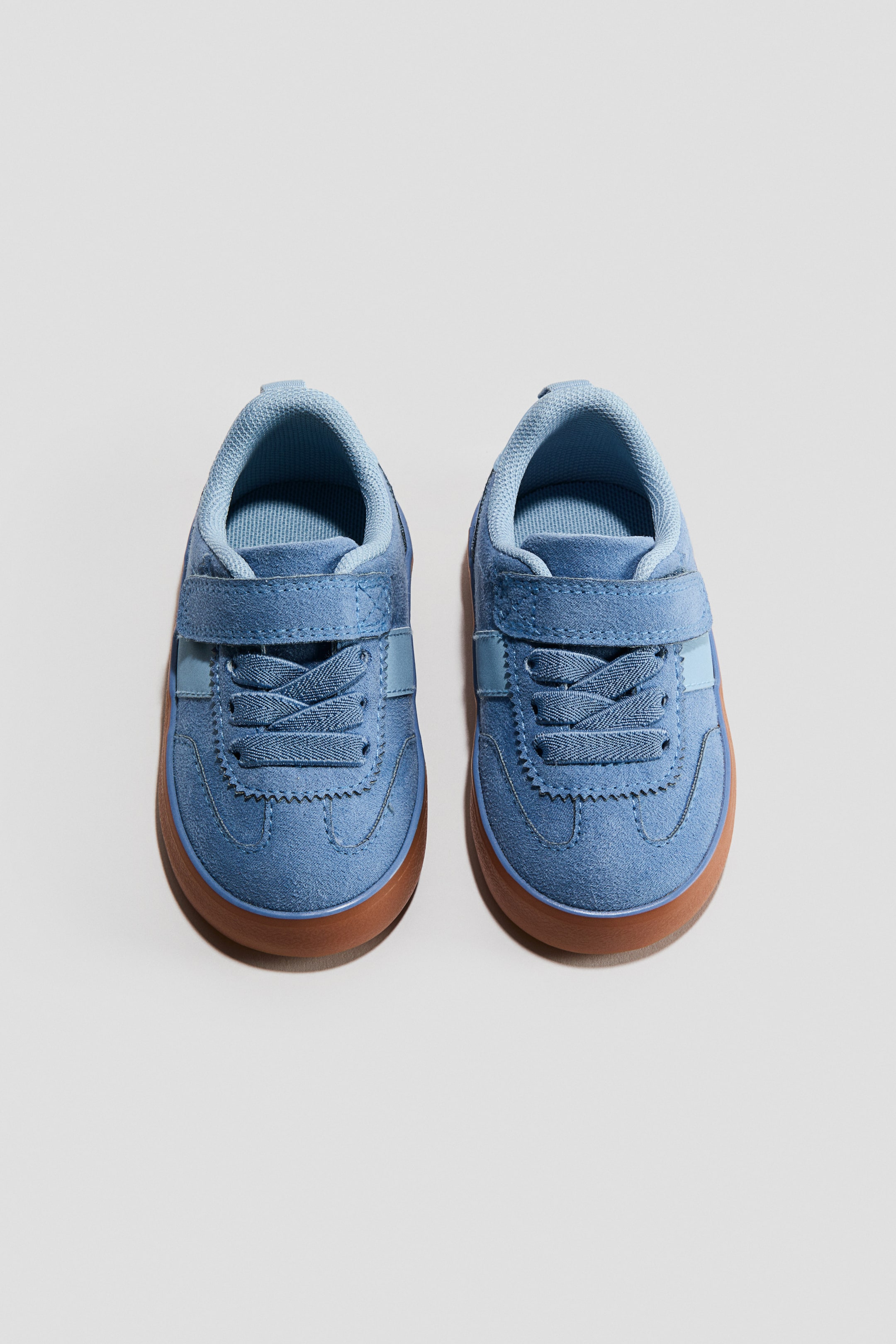 Visualizza immagine più grande: Un paio di sneakers blu a profilo basso è presentato FRONTE, dotato di una patta con chiusura hook-and-loop che si sovrappone ai lacci elasticizzati. Ciascuna scarpa presenta colletto e linguetta imbottiti, con una suola liscia marrone visibile attorno alla base.