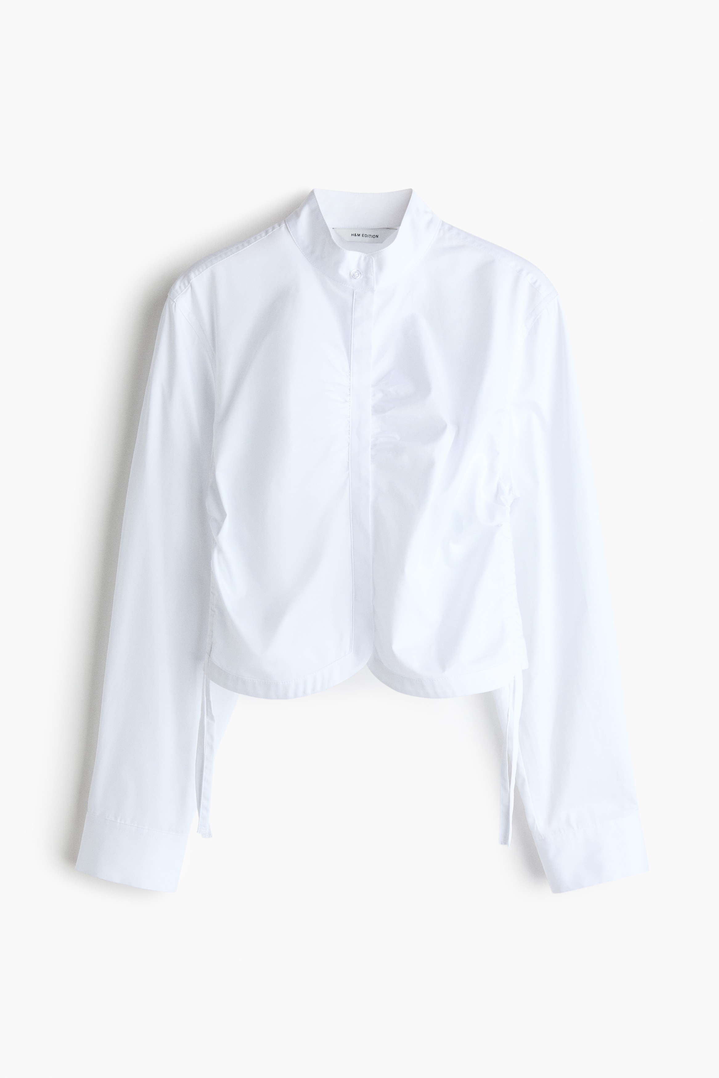 Una camicia cropped in popeline di cotone bianco Ã¨ indossata dalla modella, con maniche lunghe e polsini con bottoni, e un coulisse laterale arricciato che crea un effetto drappeggiato. La camicia Ã¨ abbinata a pantaloni in pelle nera, lasciando scoperto il lato della vita.