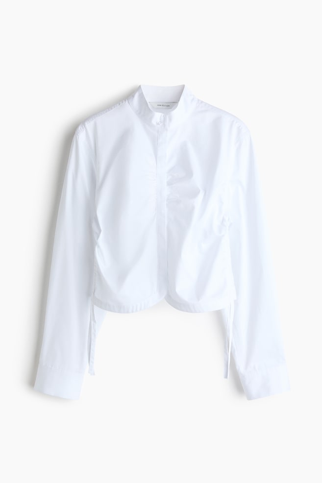 A white shirt is featuring a stand-up collar and a concealed front placket with a visible top button. It is also showcasing ruched detailing down the center front, a cropped and curved hemline with side ties, and long sleeves with buttoned cuffs. - 1