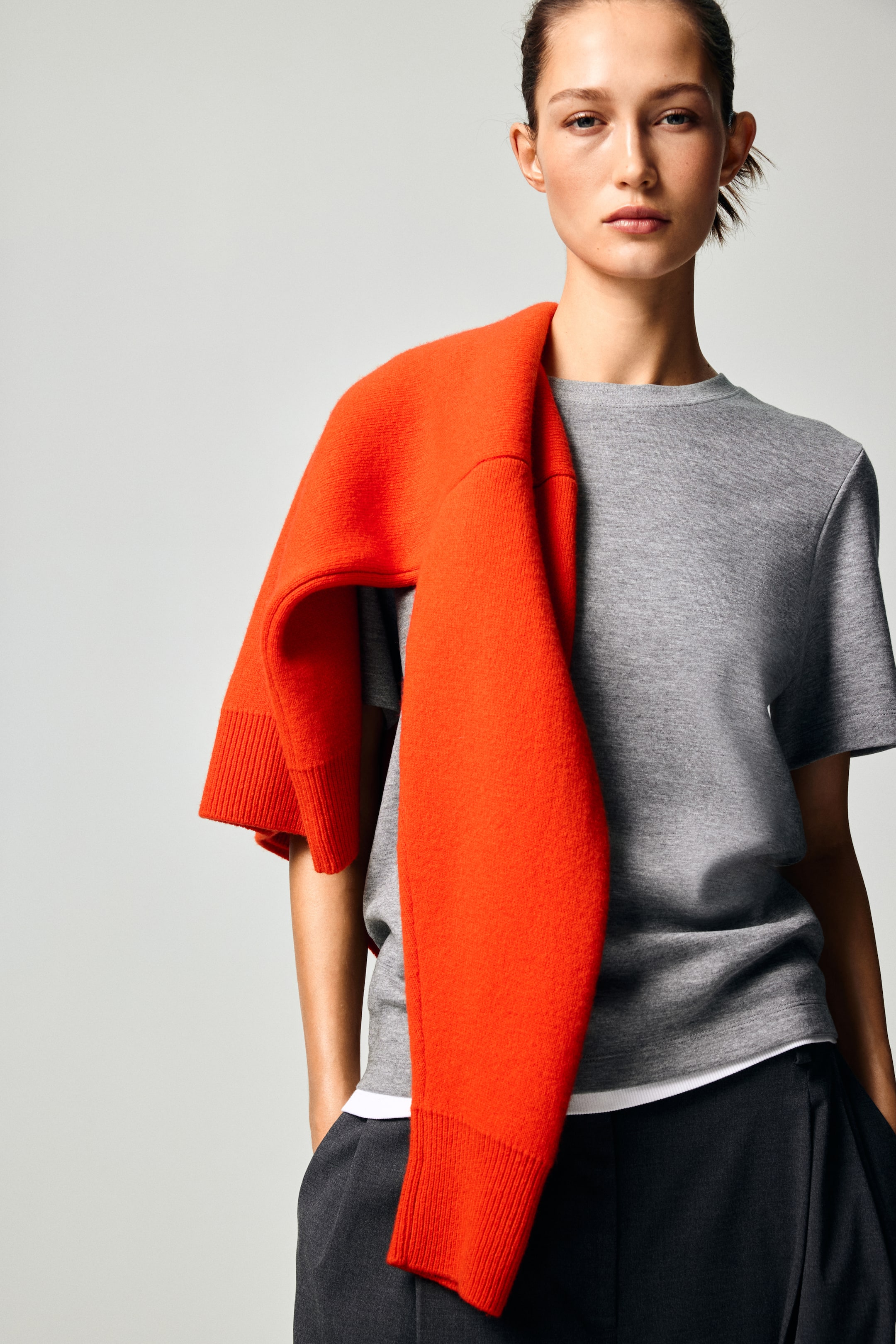 View larger image: An orange knit sweater is draped over the shoulders, its long sleeves resting across the front. The sweater features ribbed cuffs and a soft, textured finish, and is styled over a light gray crew-neck T-shirt.