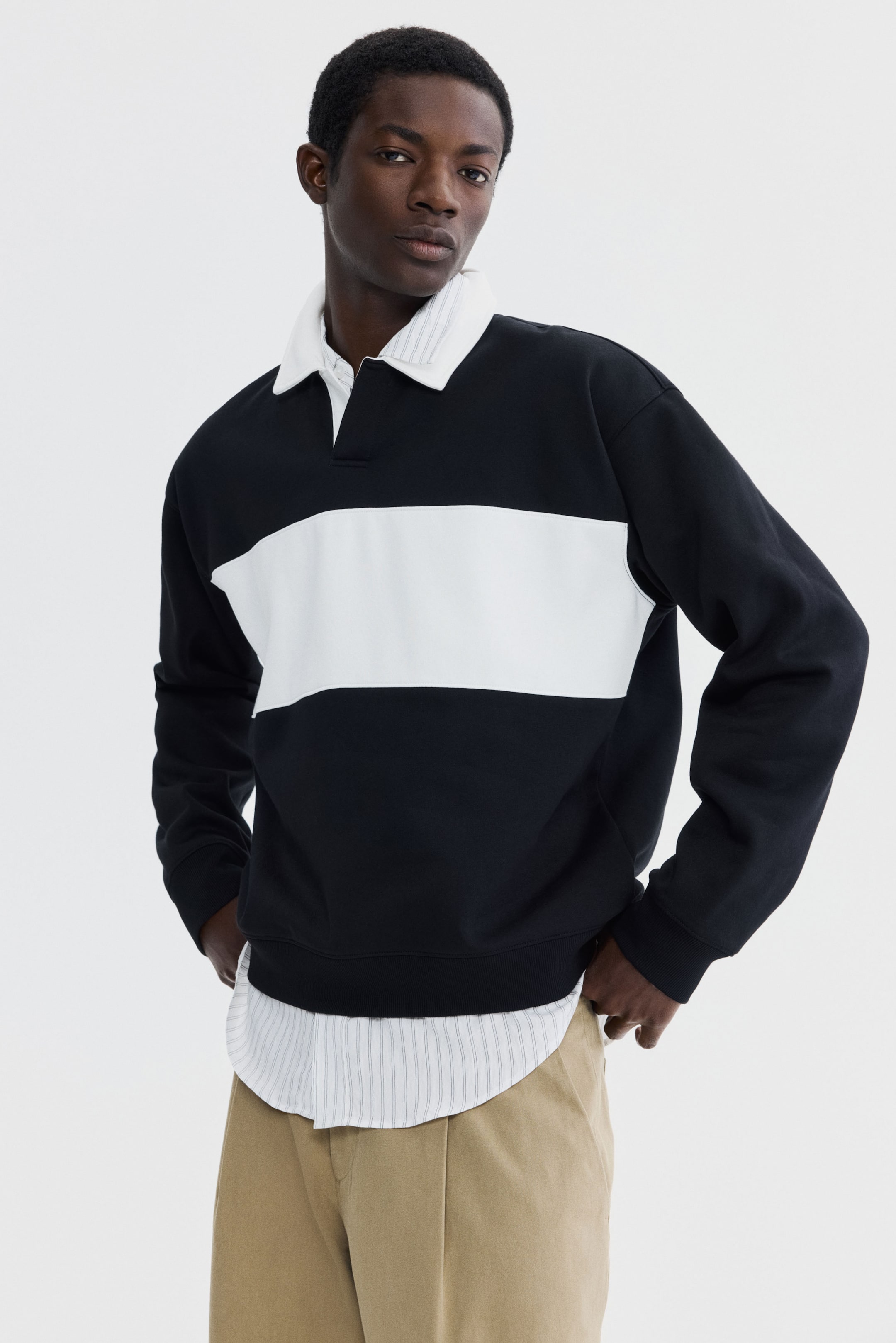 View larger image: The model is wearing a black sweatshirt top featuring a white horizontal stripe across the chest, complemented by an open white collar, dropped shoulders, long sleeves with ribbed cuffs, and a ribbed hem. Beneath the loose-fitting top, a white collared shirt is layered, adding a touch of sophistication.