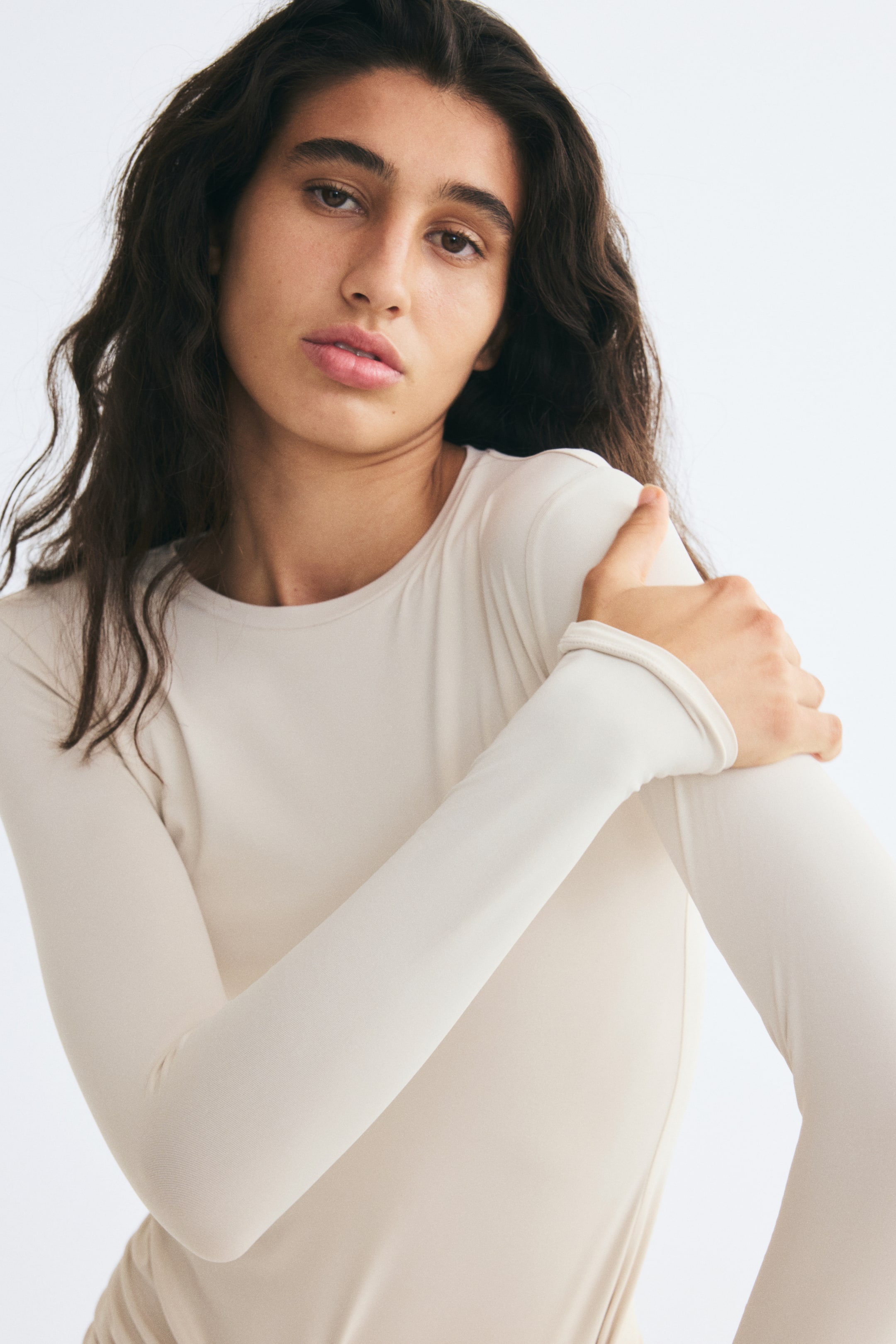 Voir une image plus grande: Un haut ajusté à manches longues beige clair avec encolure ronde est porté par le mannequin. Le mannequin tient son épaule opposée, mettant en valeur la texture jersey douce et l’ajustement près du corps de la manche.