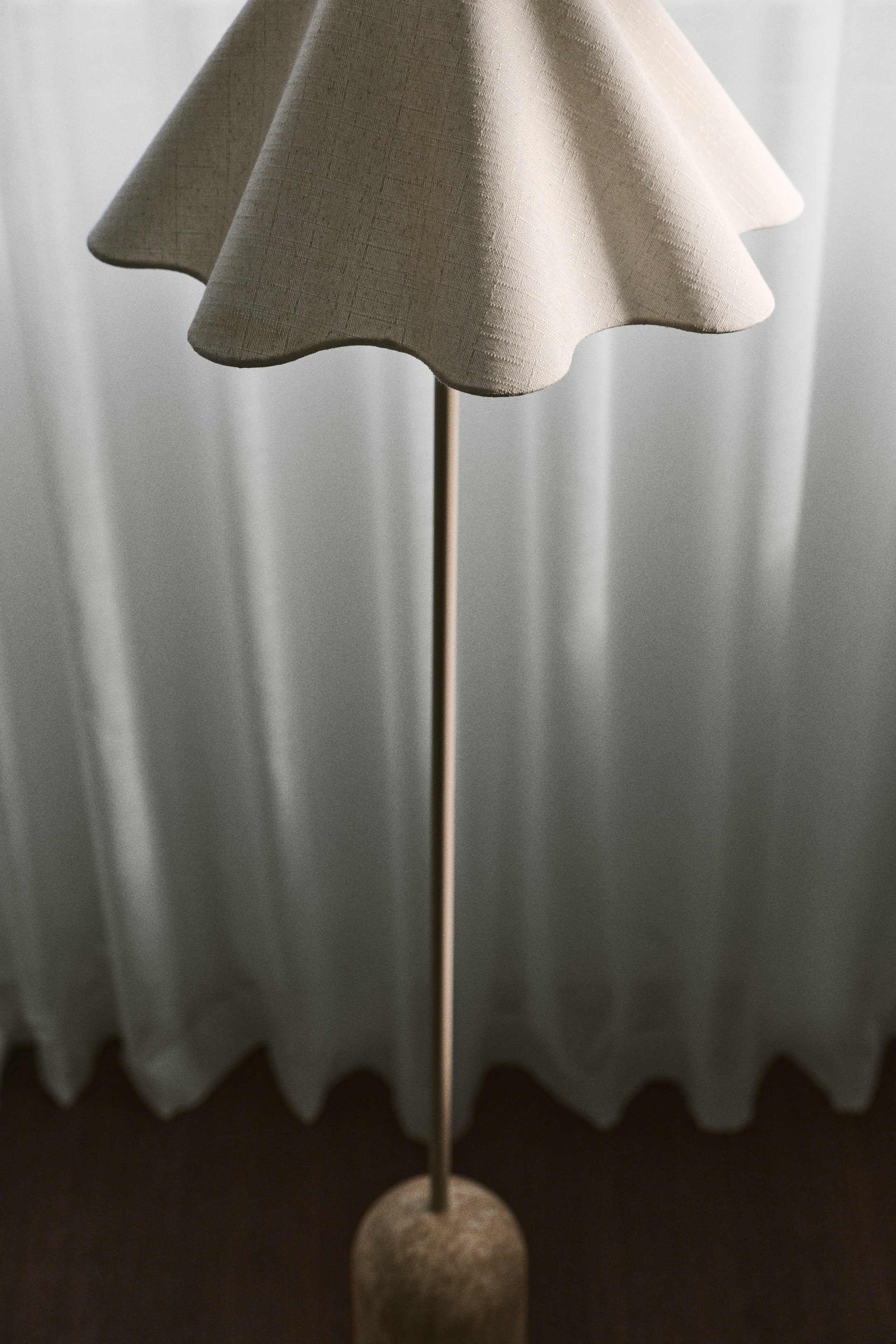 Grotere afbeelding bekijken: Een staande lamp wordt van voren afgebeeld en heeft een elegante, golvende lampenkap van beige stof. De lampenkap rust op een slanke, elegante metalen staander die uit een bedrukte cilindervormige voet omhoogkomt.