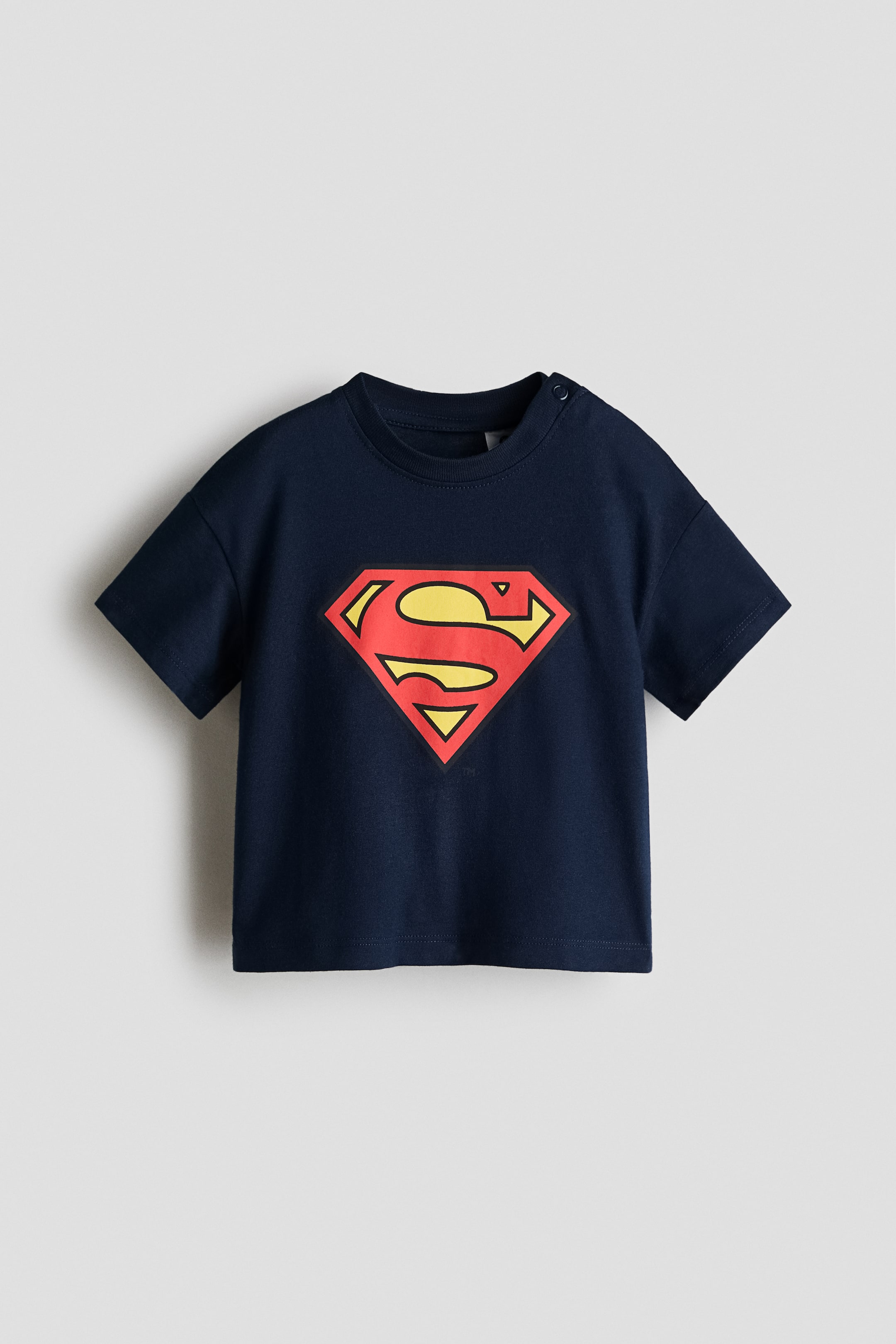 View larger image: A navy-blue T-shirt is front-facing, displaying a red-and-yellow Superman logo on the chest. It has short sleeves, with snap buttons securing one shoulder.