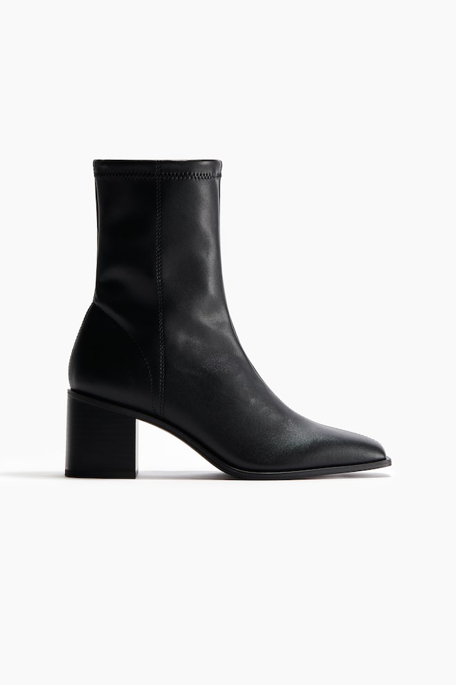 A black ankle boot is featuring a pointed toe and a stacked block heel. The boot's fitted shaft is showcasing a vertical seam along the back and a visible zigzag stitch around the top opening. - 1