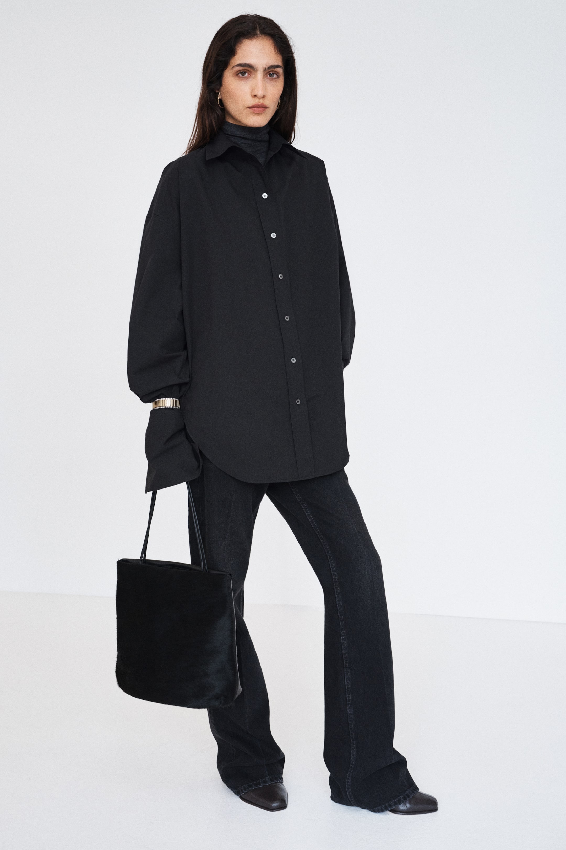 Grösseres Bild ansehen: Das Model trägt ein oversized schwarzes Hemd, kombiniert mit einem dunklen Mock-Neck-Top und dunkelgrauen Wide-Leg-Jeans. Das Hemd verfügt über einen spitzen Kragen, kontrastierende Knöpfe auf der Vorderseite, überschnittene Schultern sowie lange Ärmel mit breiten, umgeschlagenen Manschetten.