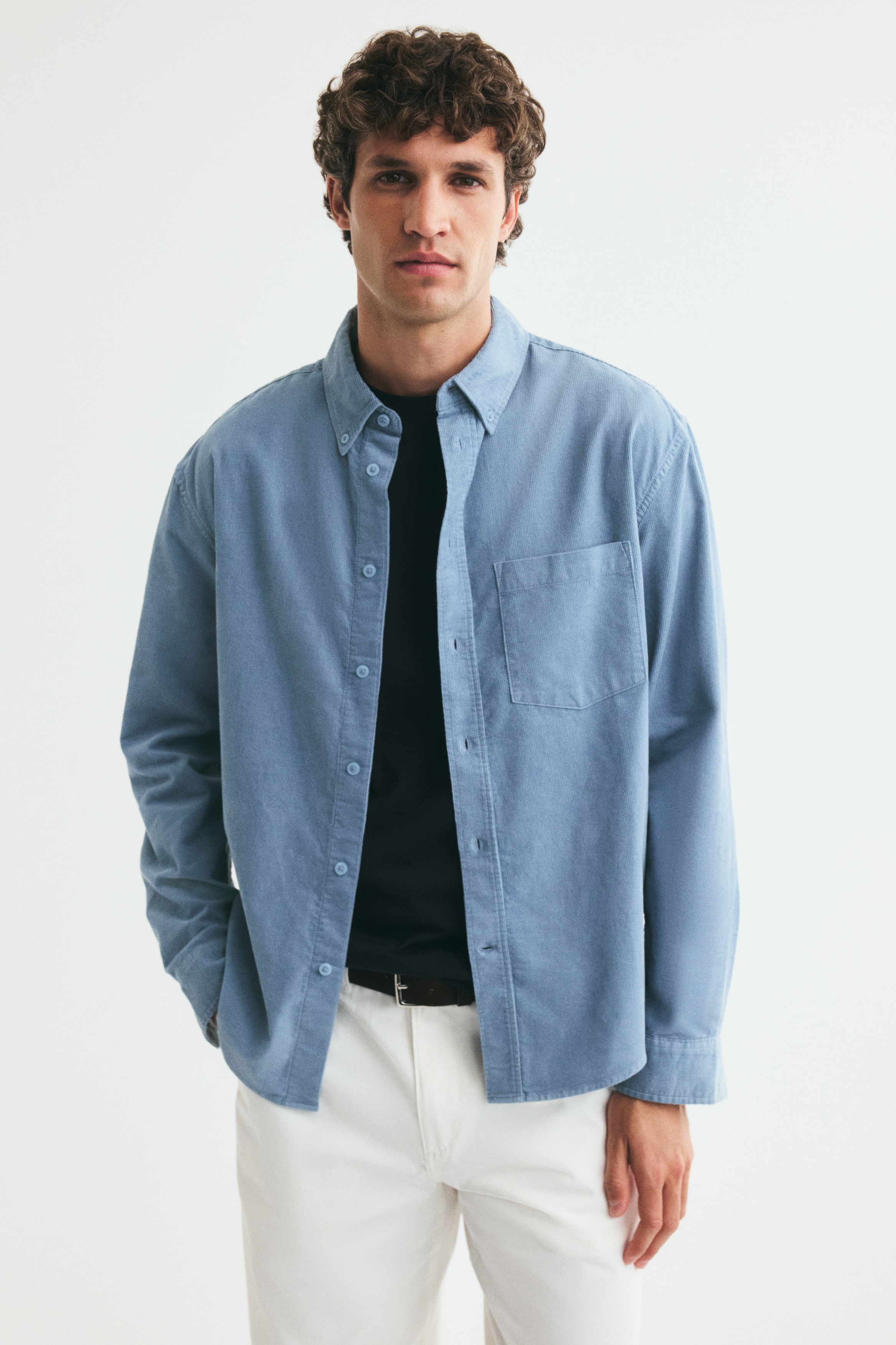 Una camisa de pana azul claro se presenta de frente, con un cuello abotonado y una tapeta completa de botones. También muestra un bolsillo parche único en el pecho izquierdo y mangas largas con puños abotonados.