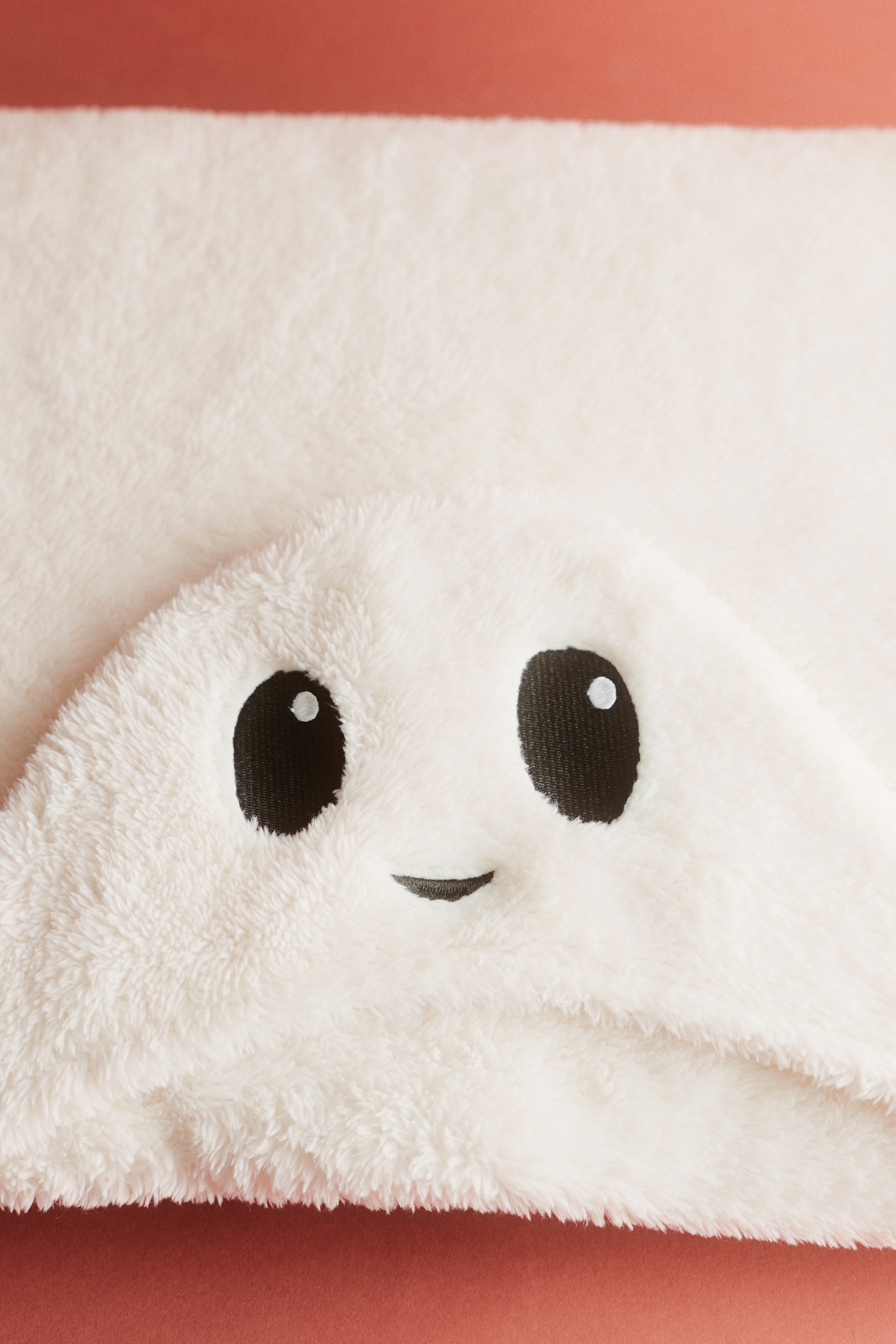 Ver imagen más grande: Una manta con capucha plegada en suave forro polar de pelo en tono blanco roto muestra detalles bordados de la cara de un personaje en la capucha orientada al frente. El rostro presenta dos grandes ojos negros con reflejos blancos y una pequeña boca negra curvada.