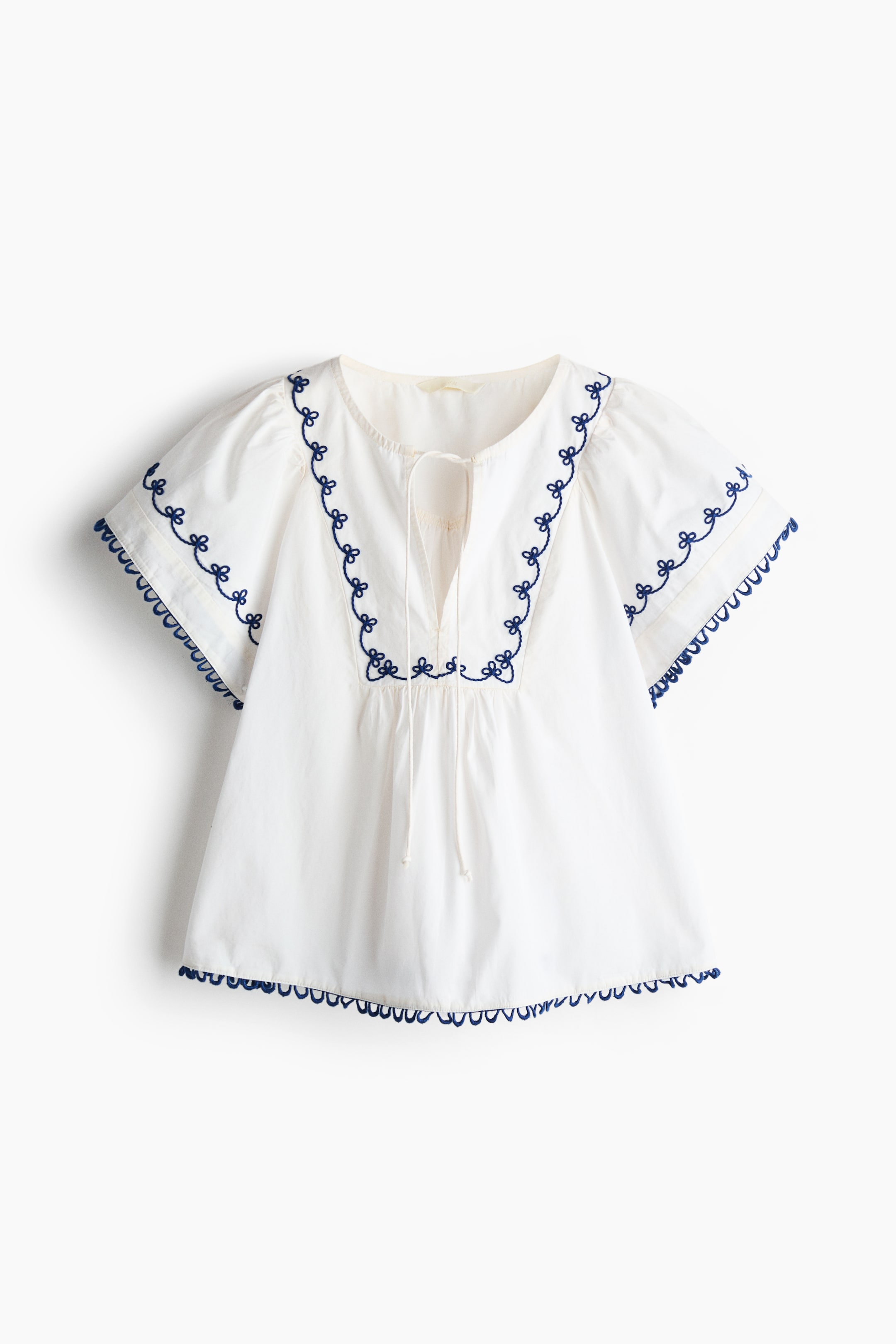 View larger image: A white short-sleeved blouse is showcasing its front, featuring a round neckline with a V-shaped opening and narrow ties. Blue embroidered floral motifs embellish the neckline and bib front with gathers, and blue picot trim decorates the cuffs and hem.