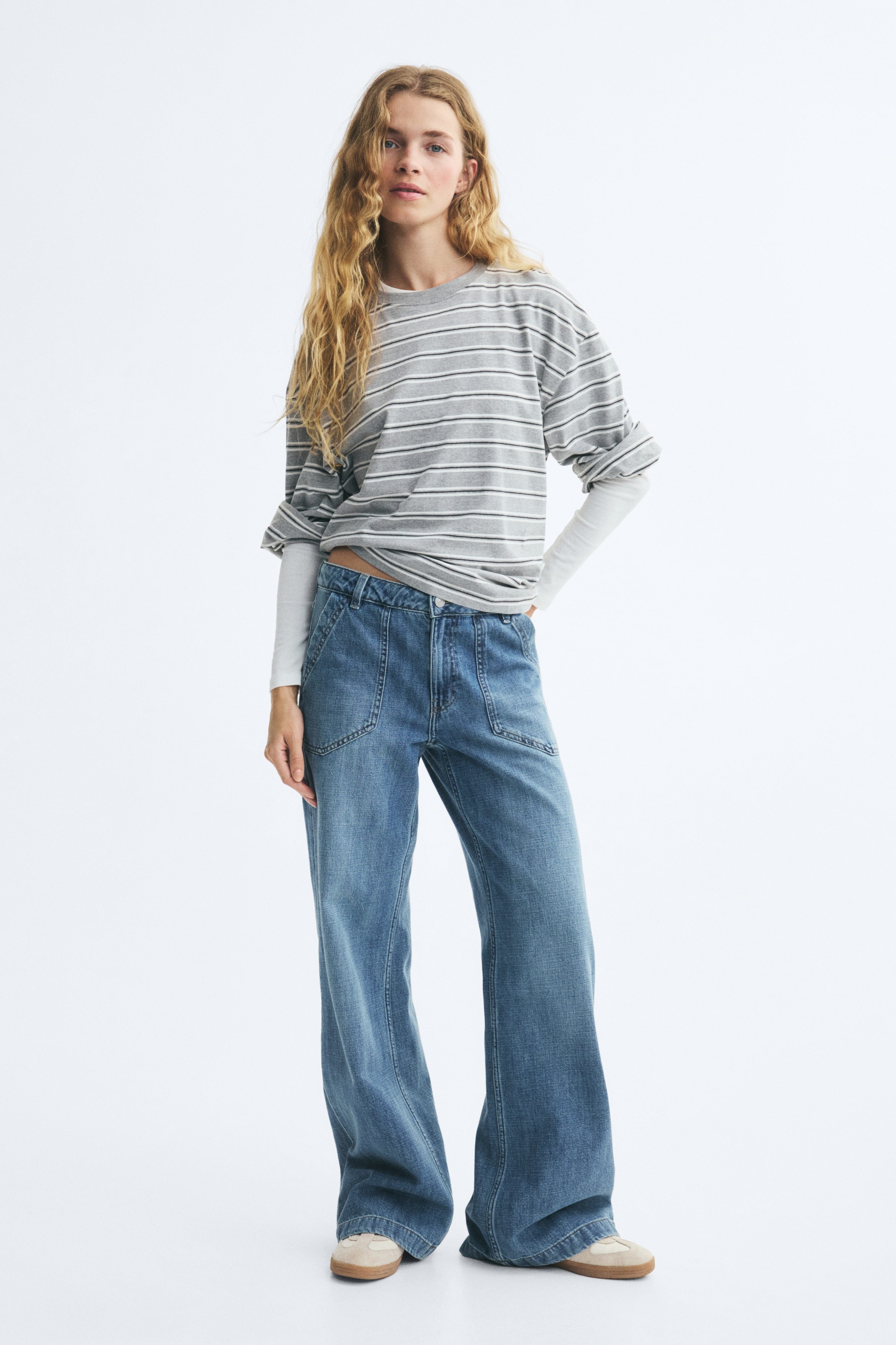 A pair of wide-leg blue jeans is shown from the front, featuring a medium-blue wash with subtle fading down the legs. The jeans showcase large front utility patch pockets, belt loops, a silver-tone button fastening at the waistband, and wide, straight-cut hems.