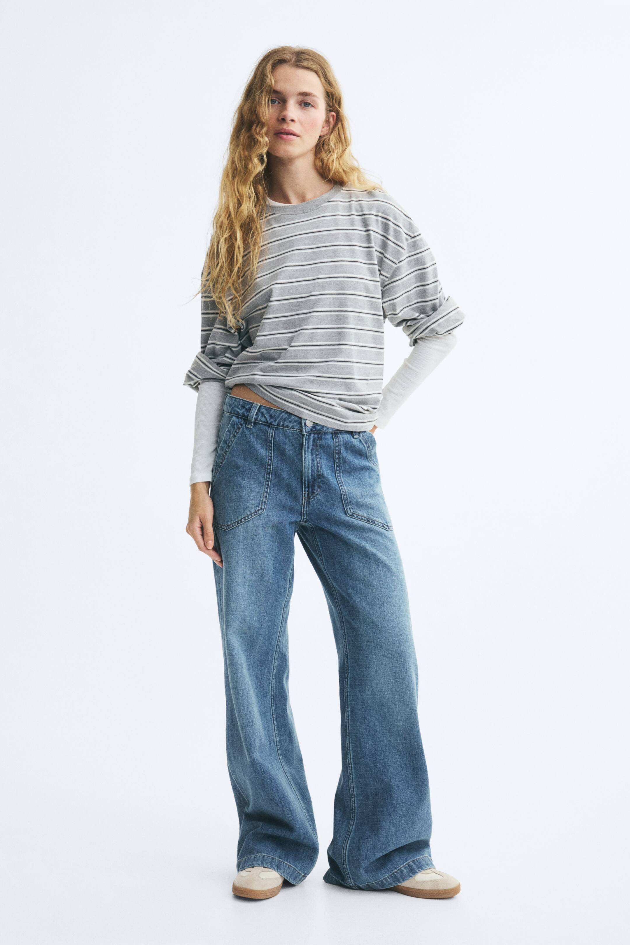 View larger image: Blue wide-leg denim jeans with a relaxed fit and prominent diagonal front pockets are being worn by the model. The jeans are paired with a layered, grey-striped t-shirt and beige sneakers.